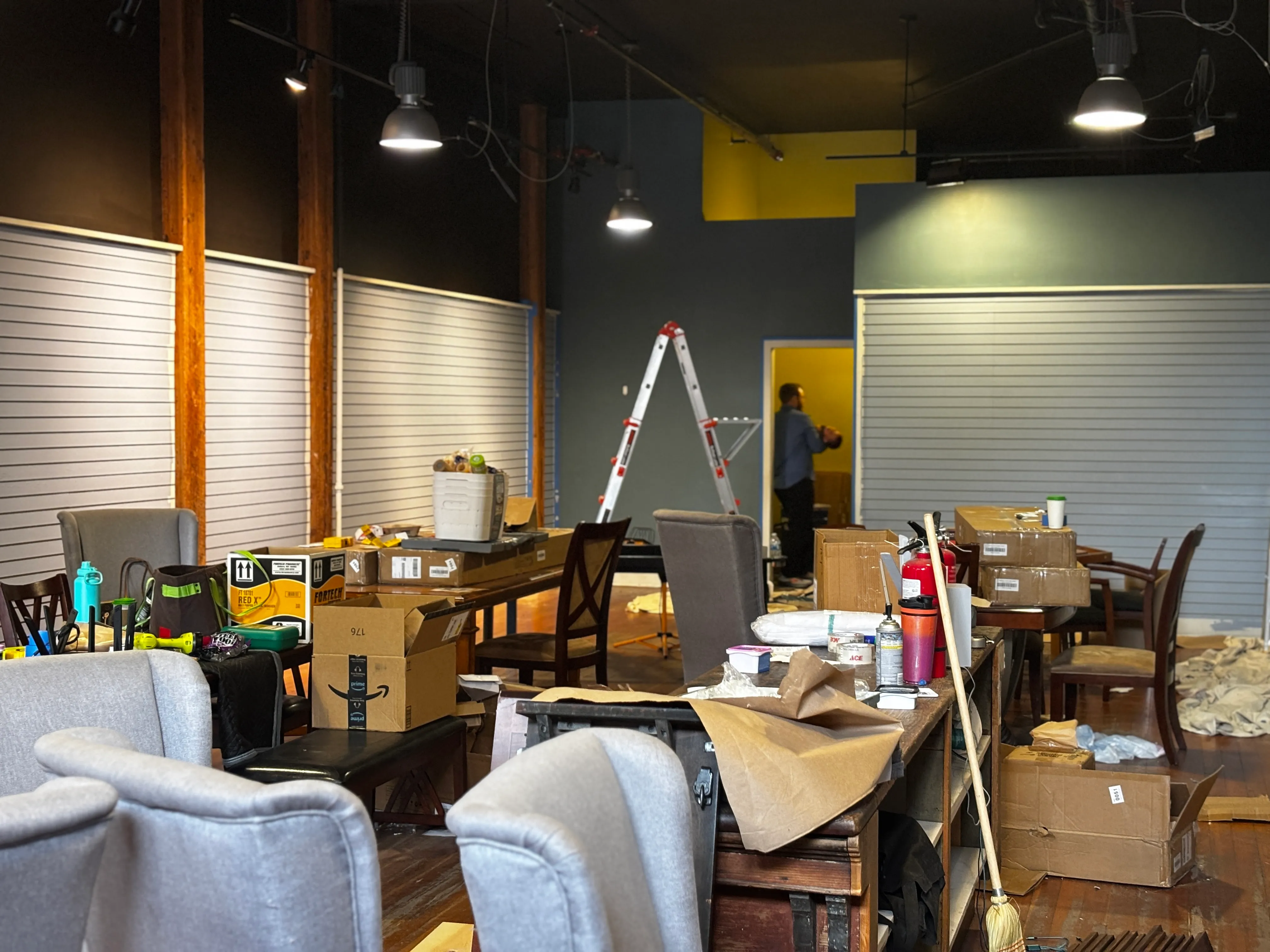 Indoor space with chairs, tables, boxes, tools, and a ladder indicating renovation or setup in progress.