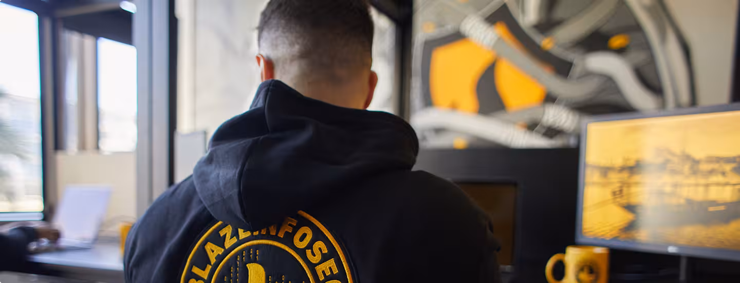 Person wearing black Blazeinfosec team member hoodie working at a desk with a monitor and mug.