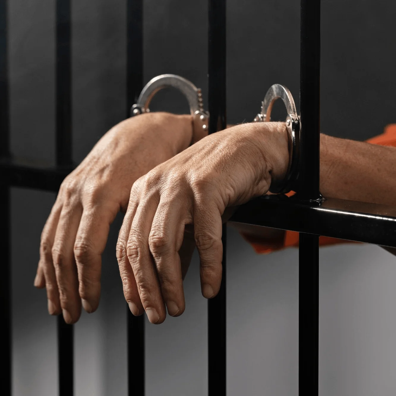 A pair of handcuffed hands rests against the bars of a jail cell, with an inmate's orange jumpsuit visible in the background.