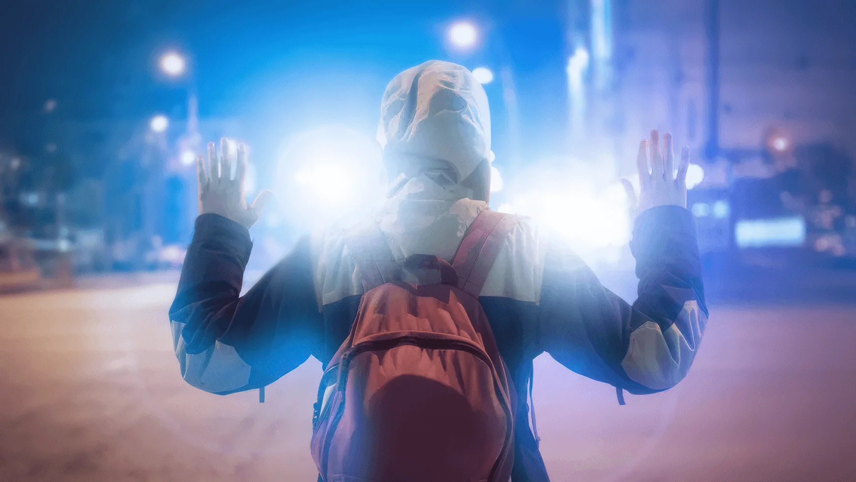 A hooded person wearing a backpack holds both hands up in front of intense police lights at night.