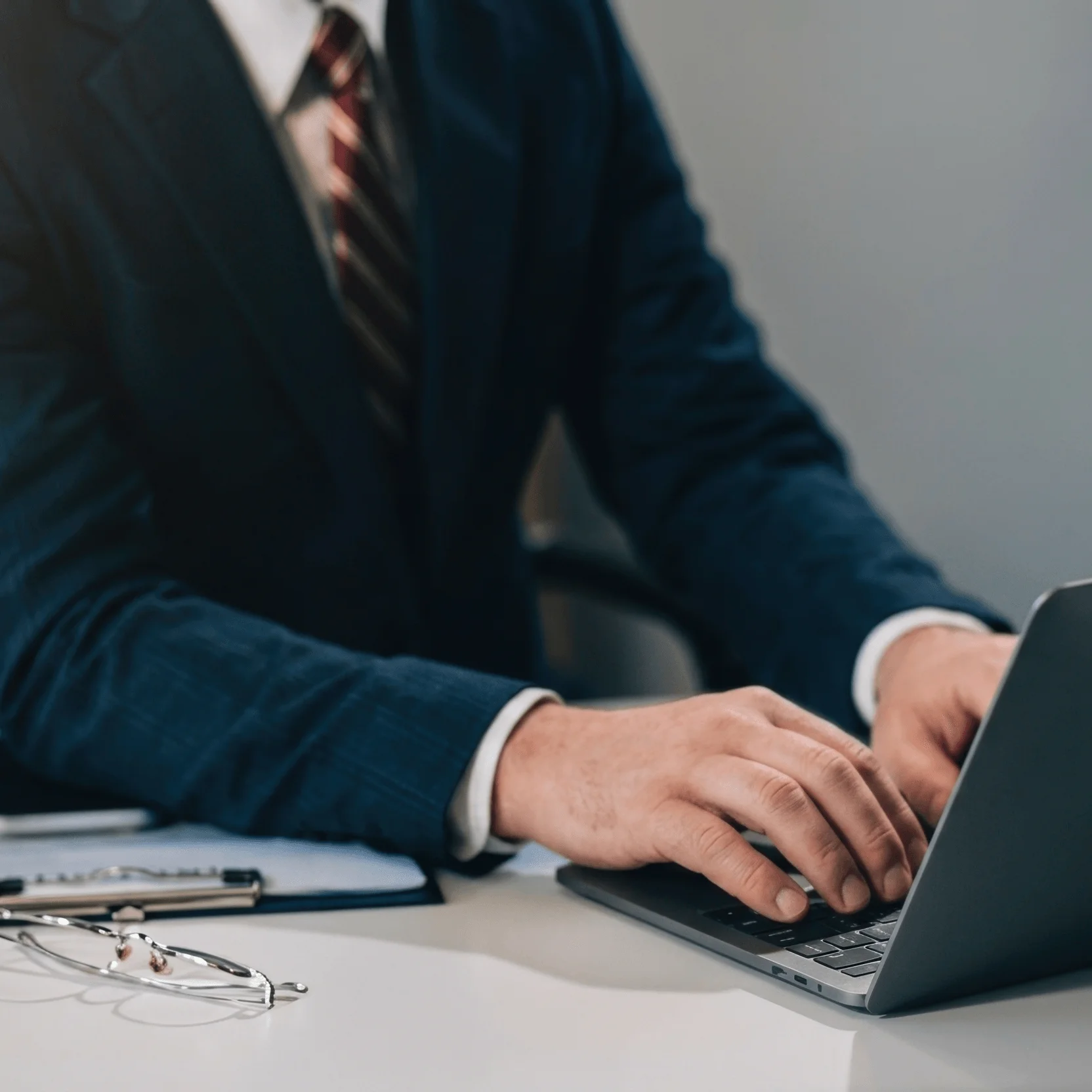 An attorney in a suit types on a laptop at his desk, with reading glasses and a legal pad resting nearby.