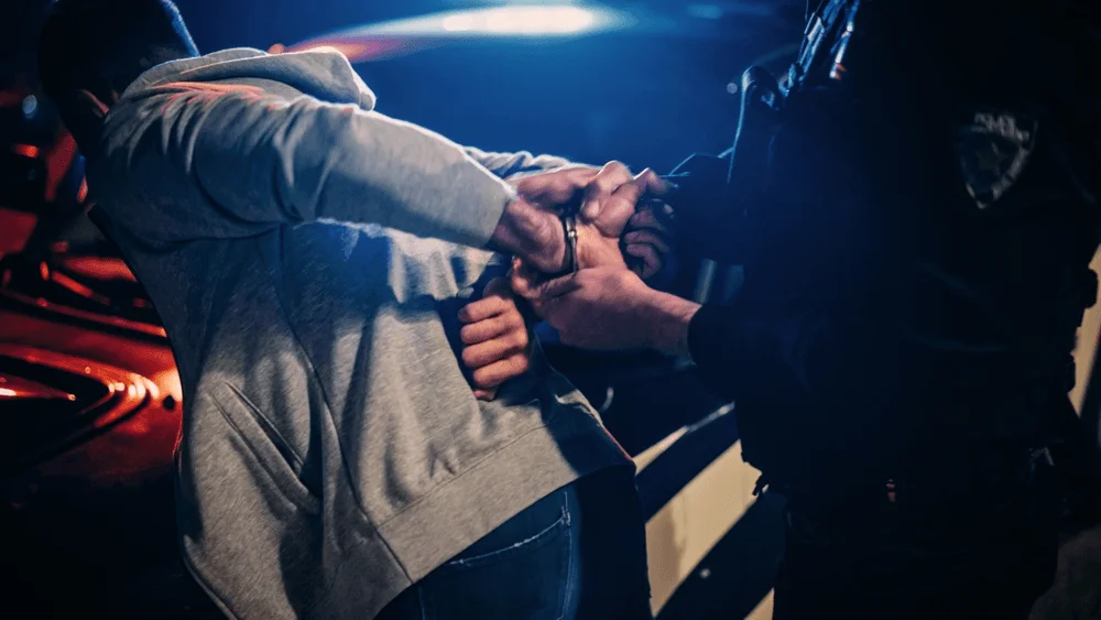 A police officer handcuffs a man at night, flashing lights illuminating the scene behind them.