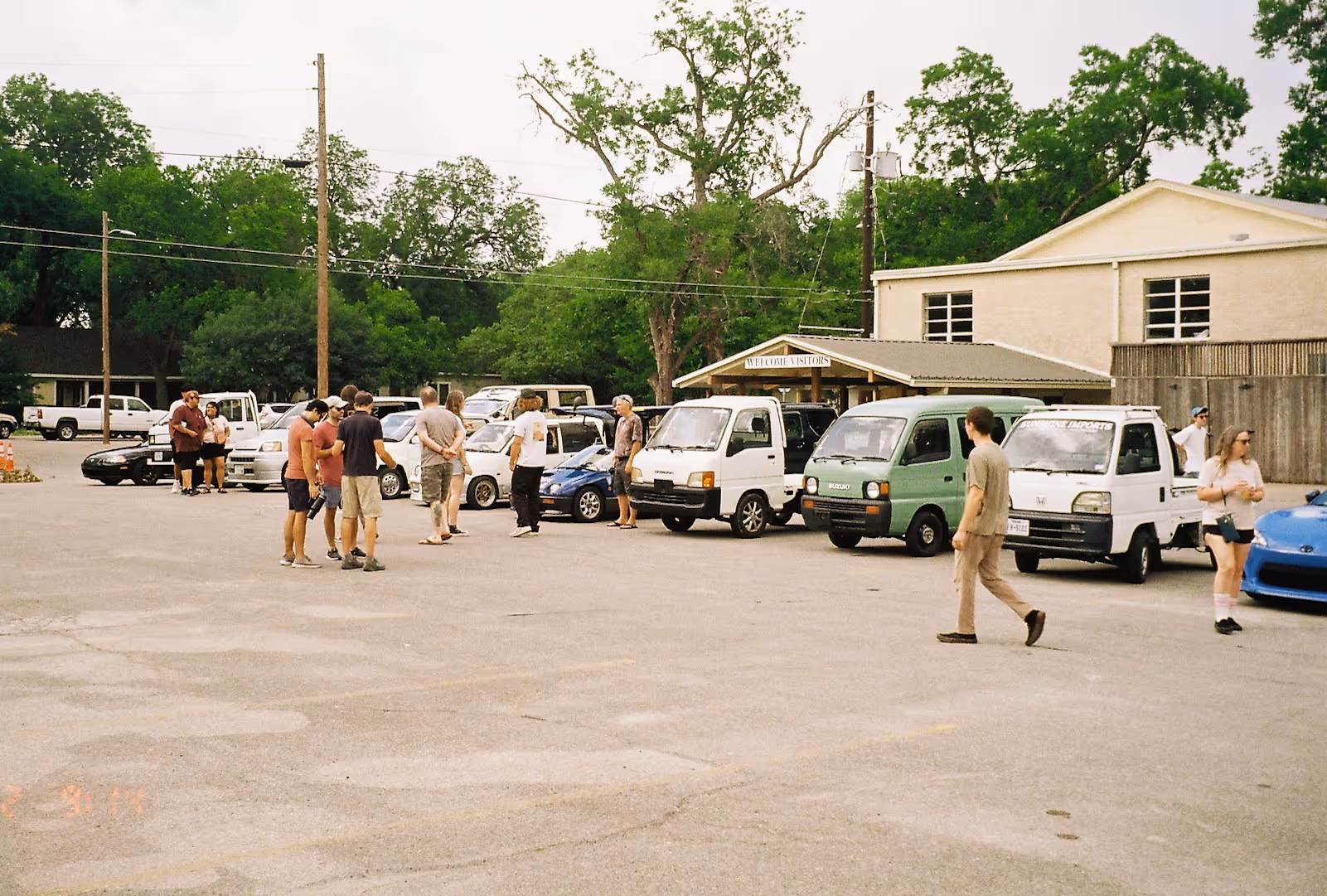 Group of people gathered in a parking lot near multiple compact cars and mini trucks with trees and buildings in the background.