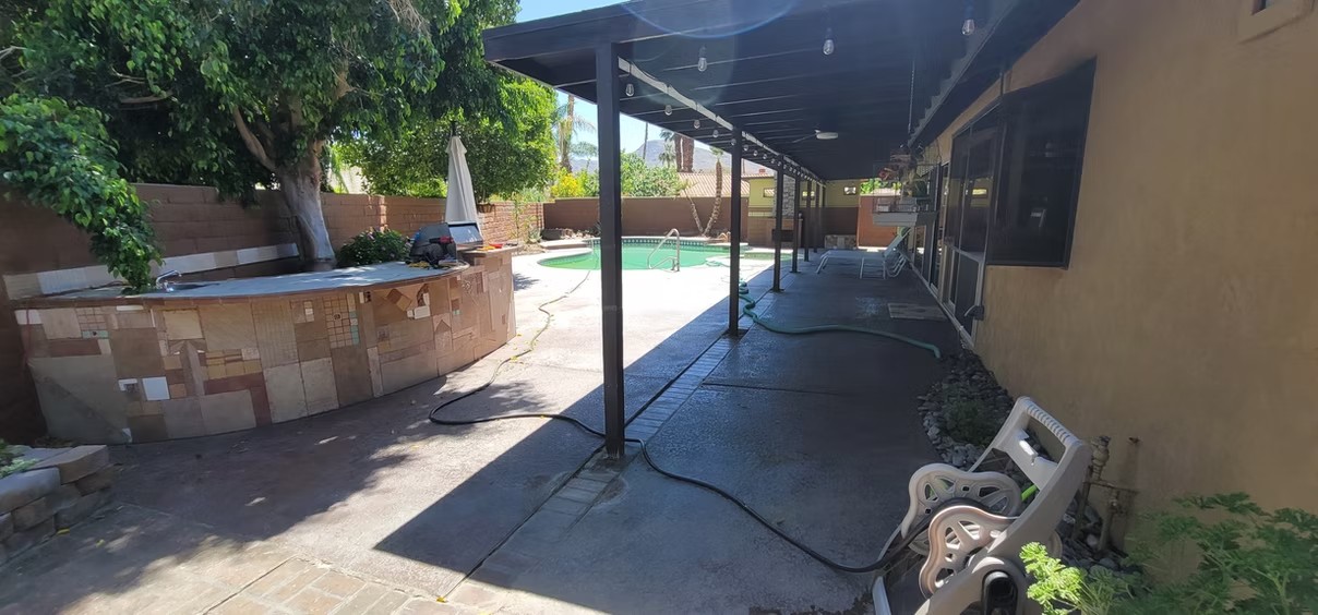 Covered patio beside a pool with tiled bar, outdoor chairs, and string lights under a roof.