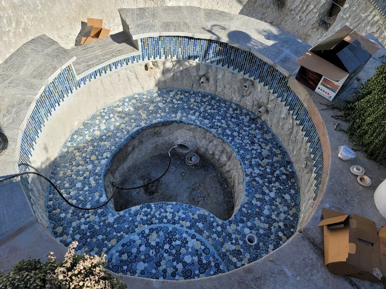 A circular spa under construction with blue pebble mosaic tiles and gray stone steps around it.