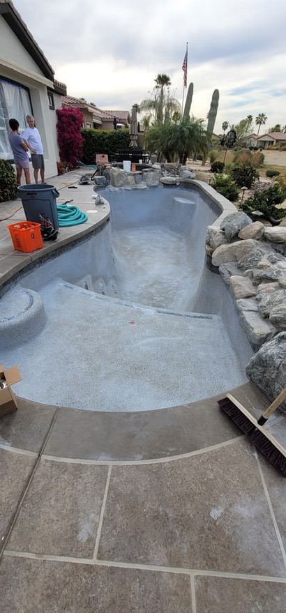 Empty residential pool under renovation with plastered surface, surrounded by stone features and backyard plants.