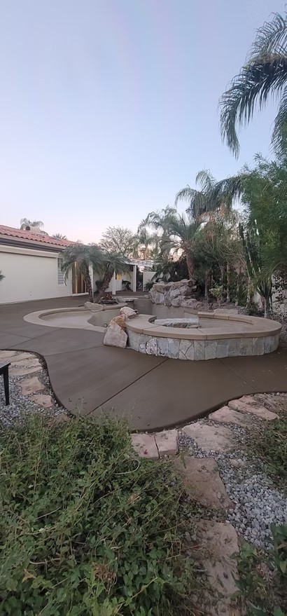 Backyard pool deck with stone fire pit, surrounded by palm trees and near a single-story house.