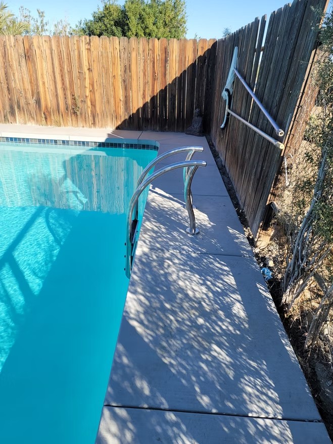 Clear blue pool with new metal handrail and wooden fence casting shadows on deck.