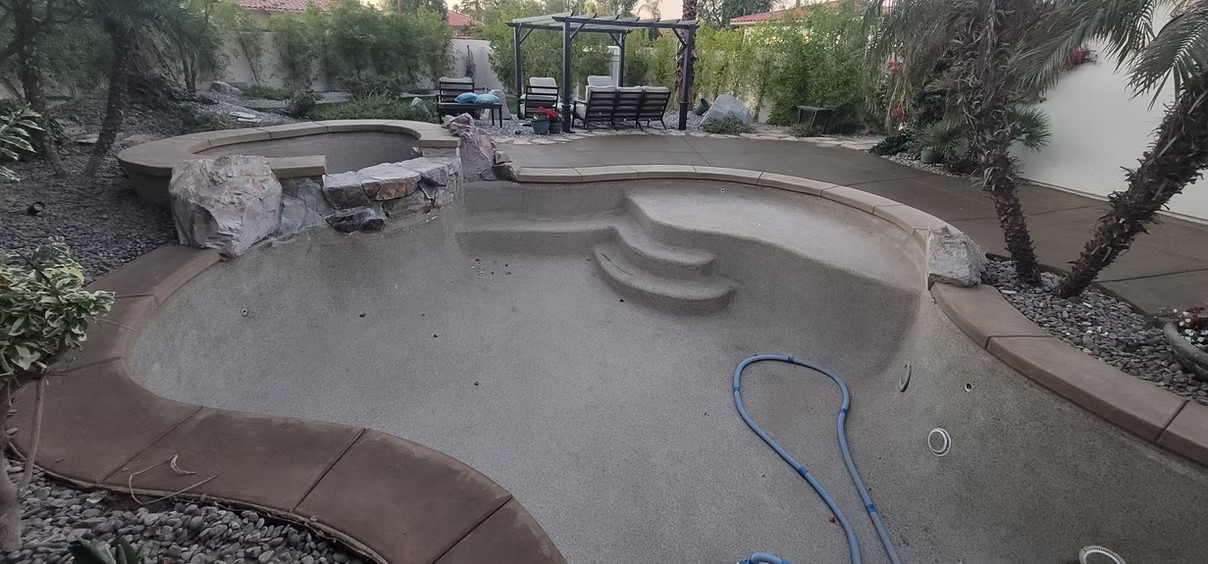 Empty backyard pool and spa under construction with stone waterfall and outdoor seating area.