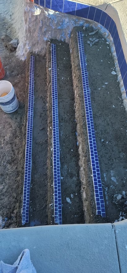 Three parallel strips of blue mosaic tiles partially embedded in a dirt trench next to a pool edge.
