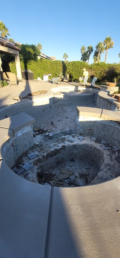 Empty pool under renovation with broken tiles and two workers removing debris near a hedge and palm trees.