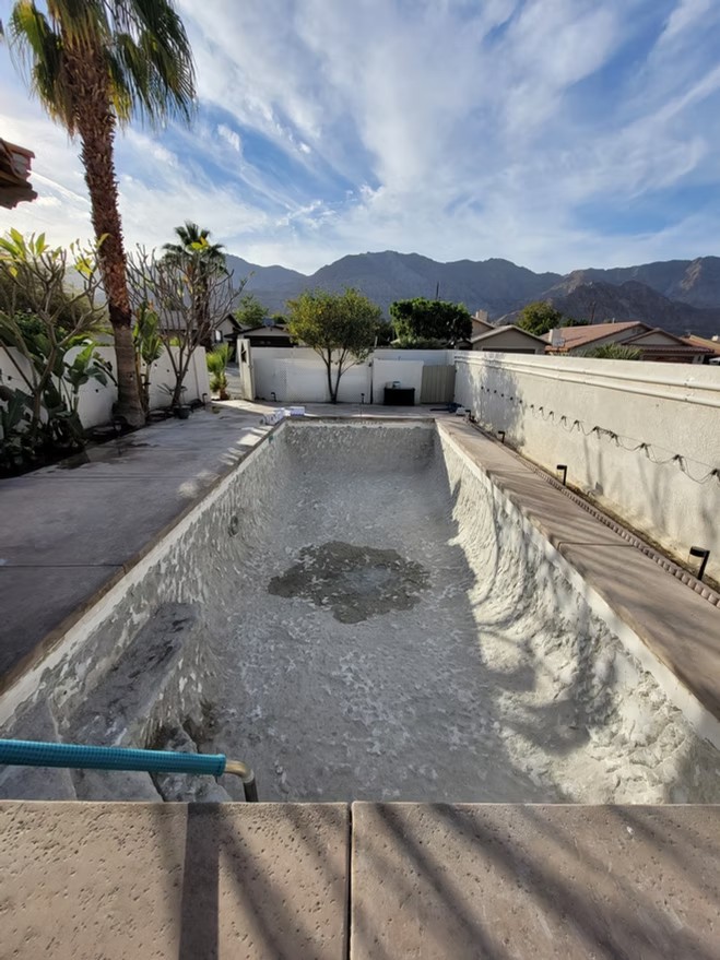 Empty residential swimming pool with chipped plaster surface under a partly cloudy sky and mountains.