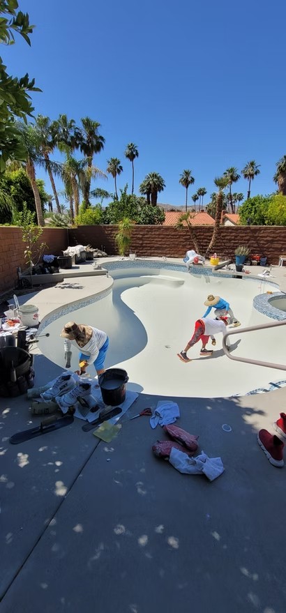 Crew plastering a white freeform backyard pool under a clear blue sky with palm trees around.