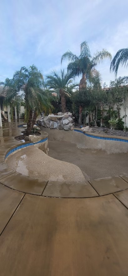 Empty backyard pool under construction with rock water feature and palm trees around it.