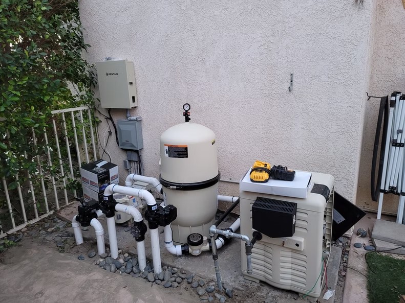 Outdoor pool equipment setup with filter tank, pump, heater, and connected PVC pipes near a house wall.