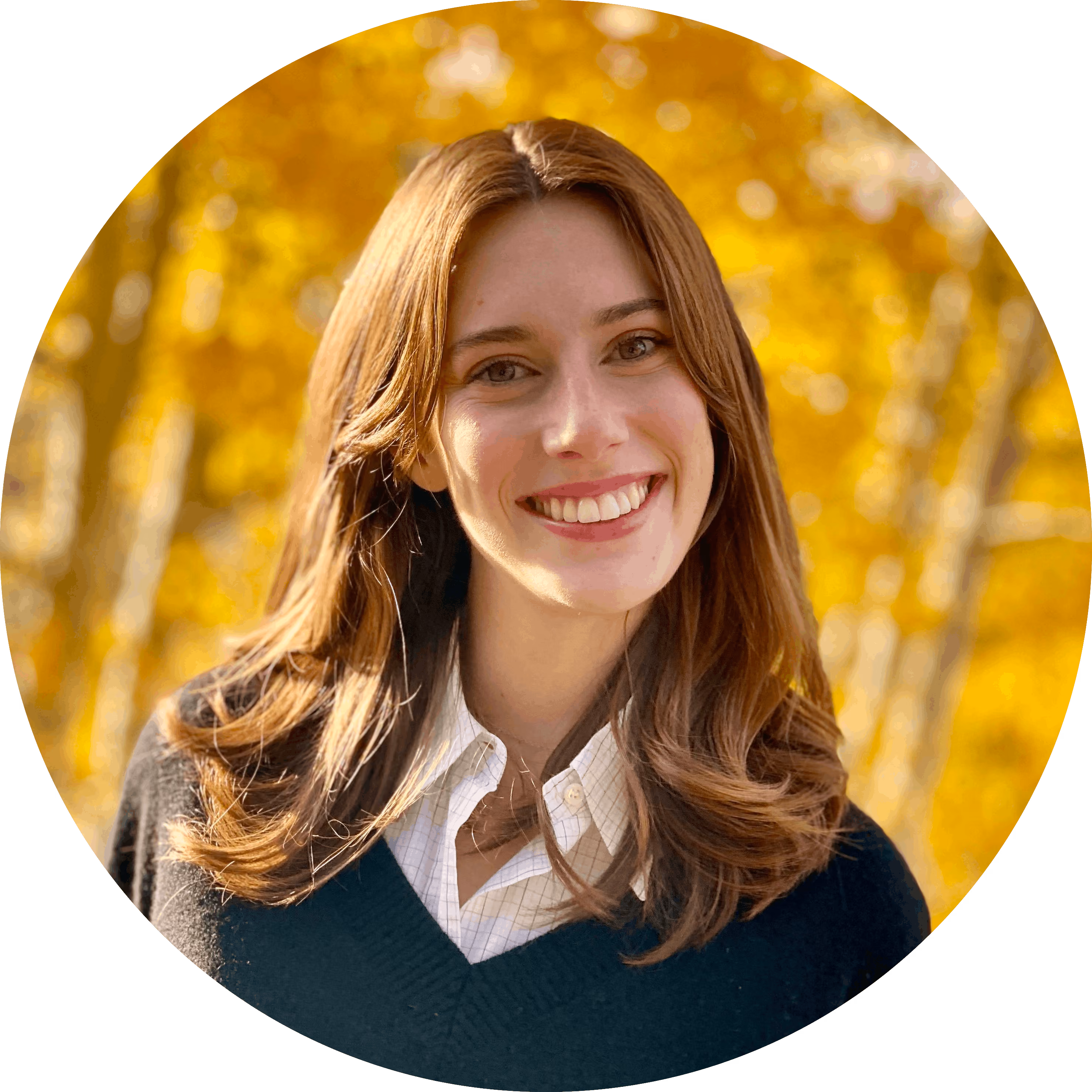 Smiling young woman with brown hair, wearing a white shirt and black sweater outdoors with fall foliage.