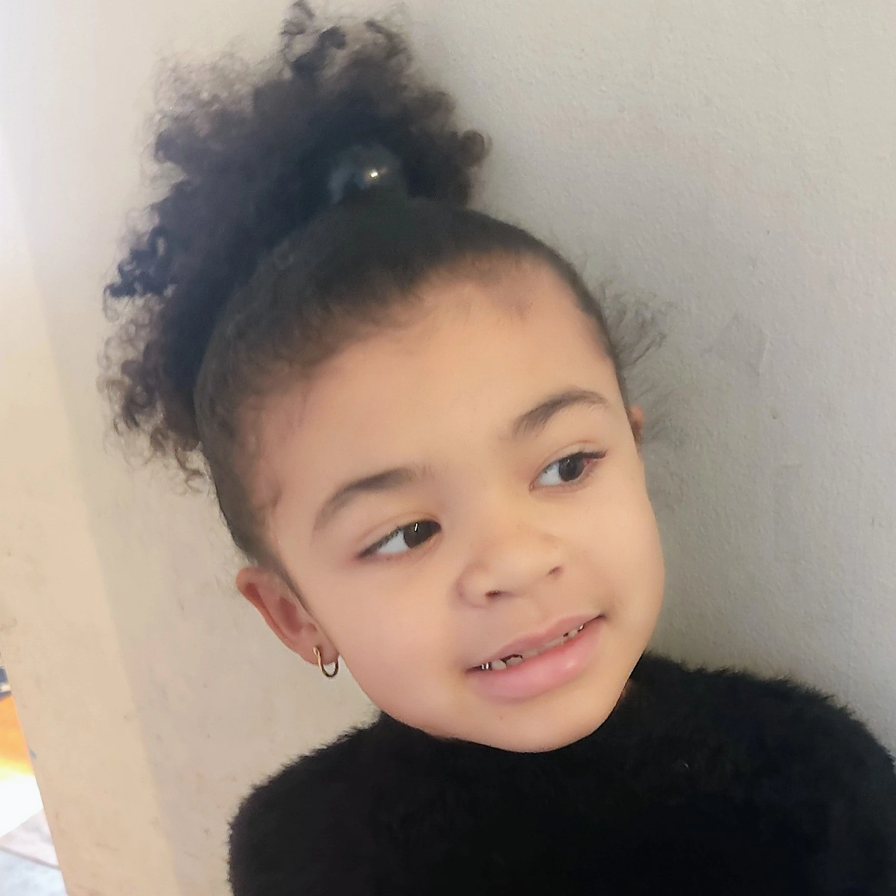 Young girl with curly hair tied up wearing small hoop earrings and a black fuzzy sweater, looking sideways.