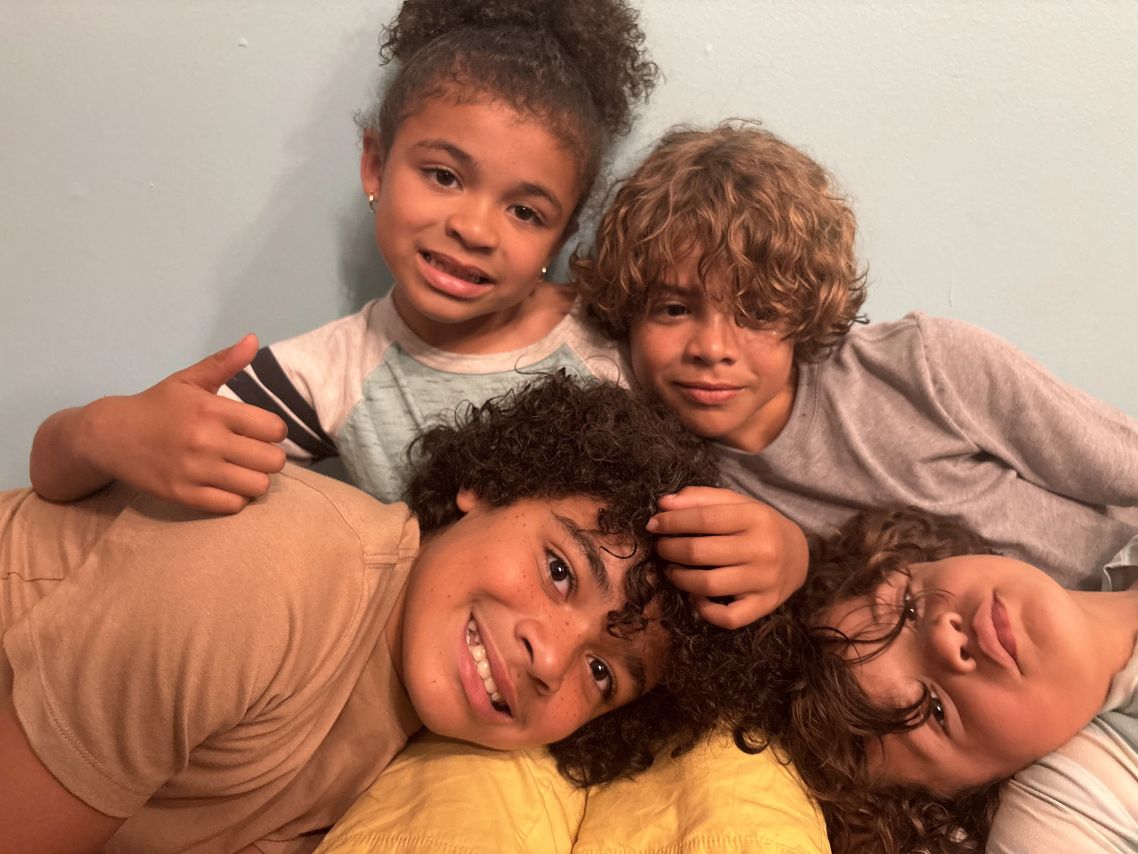 Four children with curly hair lying close together against a light blue wall, smiling and relaxed.