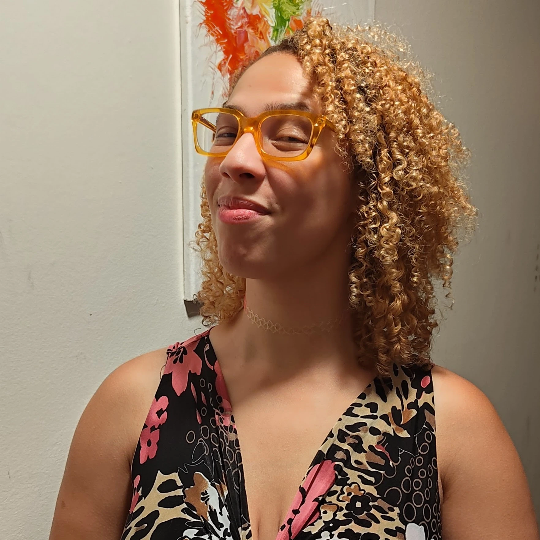 Woman with curly blonde hair wearing yellow glasses and a floral patterned dress smiling indoors.