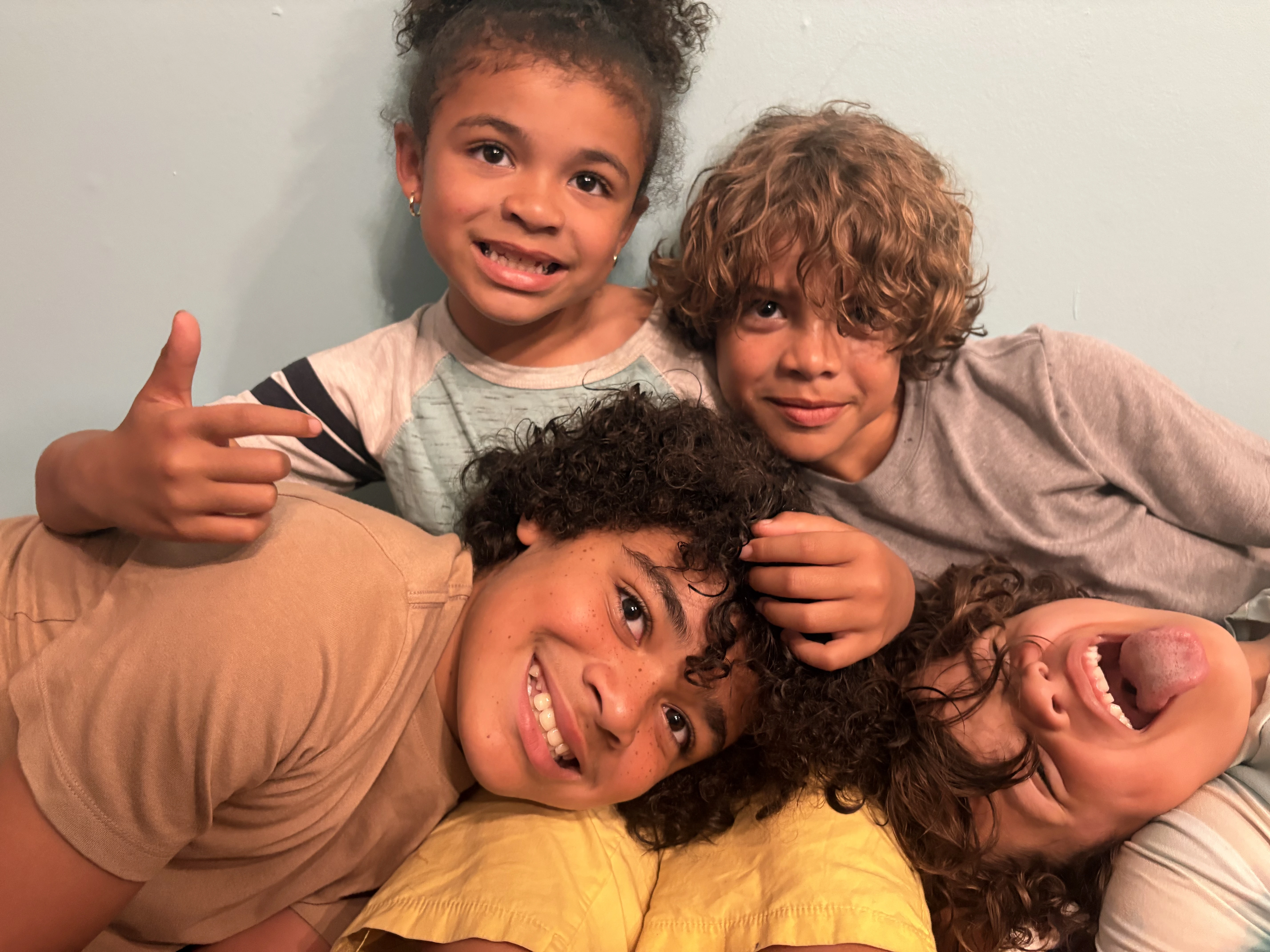 Four children with curly hair smiling and laughing close together against a light blue wall.