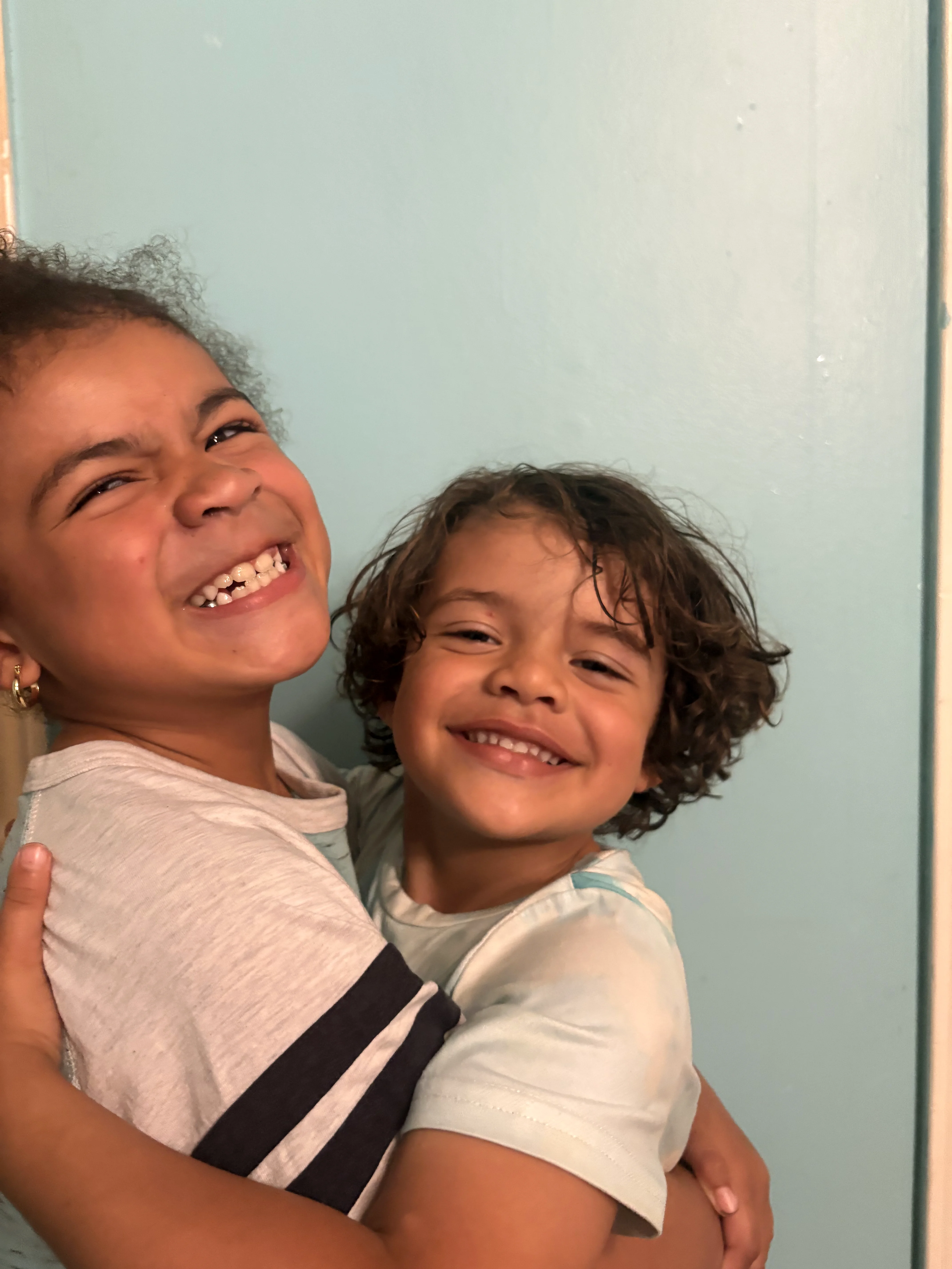 Two curly-haired kiddos hugging and smiling against a light blue wall