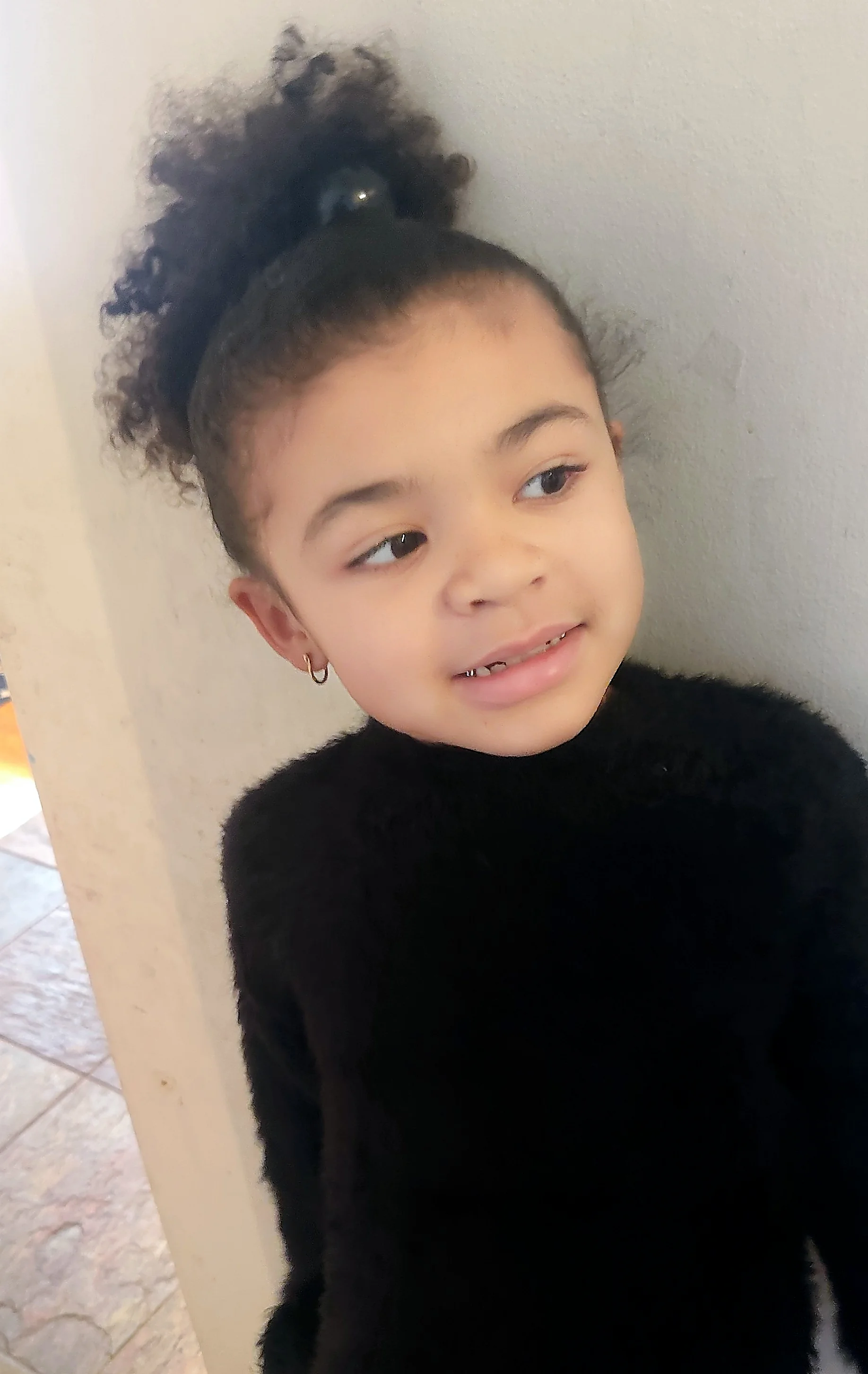 A young child with curly dark hair styled in an updo with a hair clip, wearing a black fuzzy sweater and small hoop earrings, smiling at the camera.