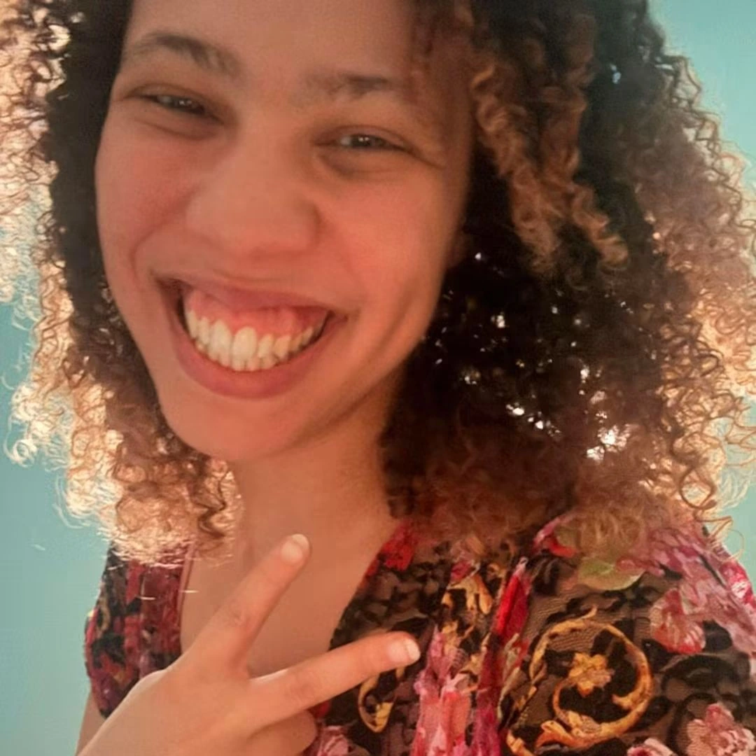 Smiling person with curly hair flashing a peace sign wearing a floral patterned top.