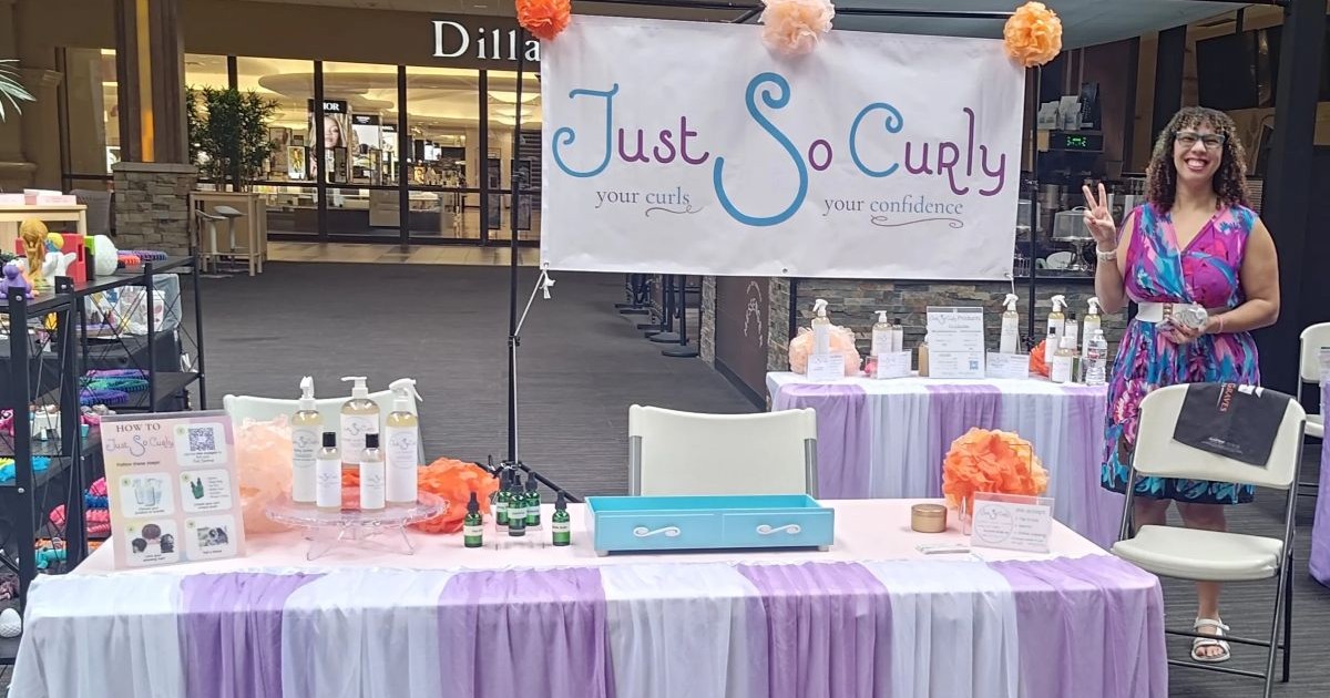 The Just So Curly pop-up table at Oak Park Mall, with the founder waving beside a 'your curls, your confidence' banner