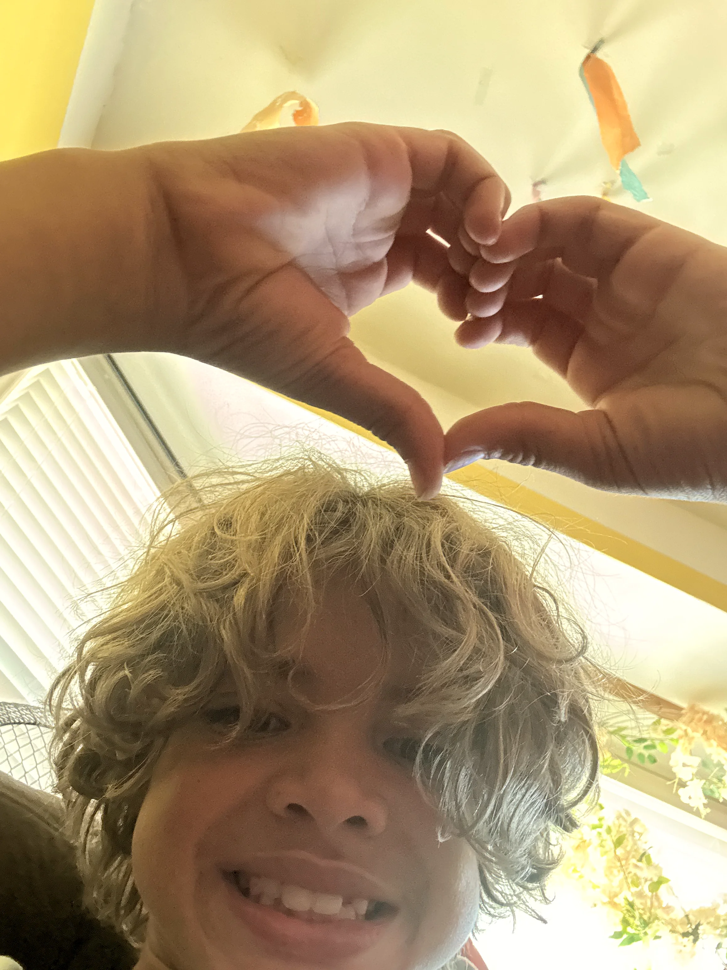 A child with curly blonde hair smiles at the camera while two hands form a heart shape above their head.