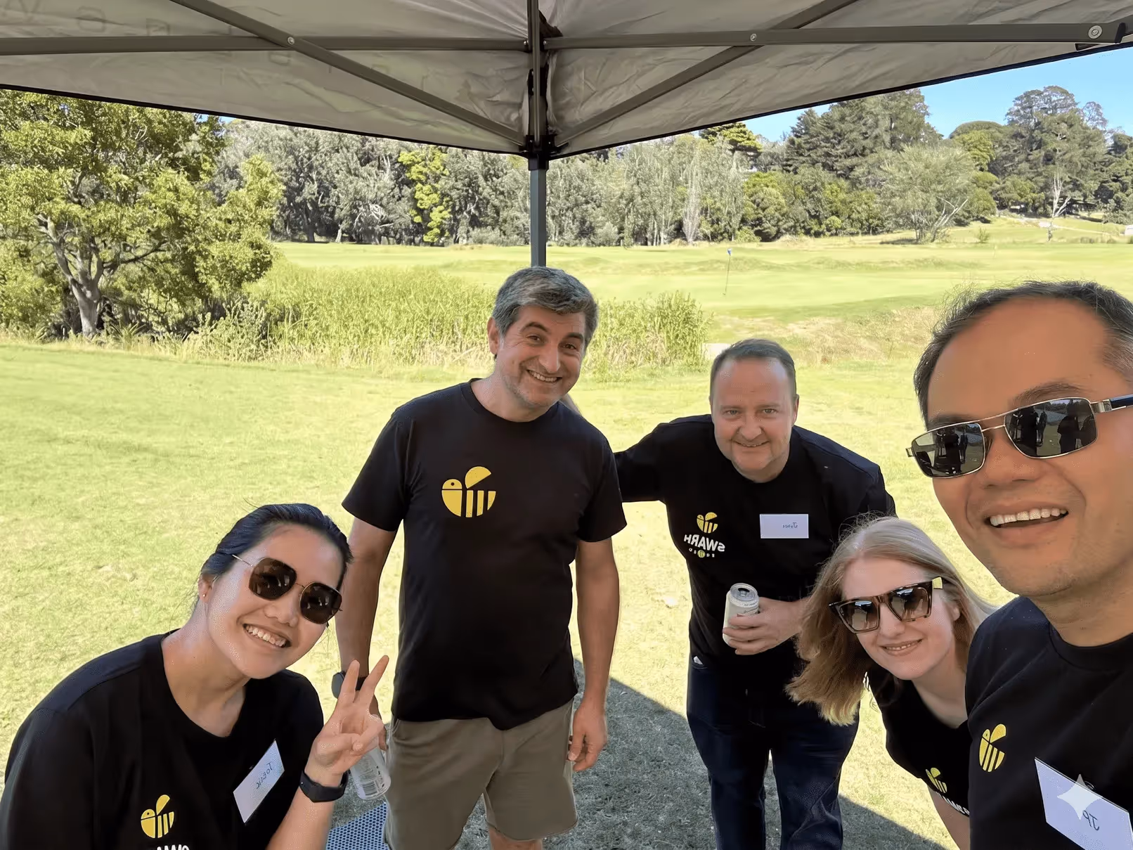 Group of five people wearing black shirts with yellow logos smiling under a canopy outdoors on grass.