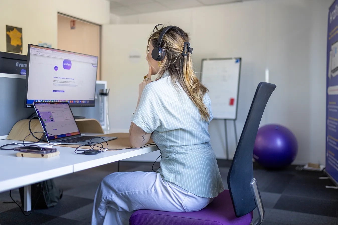 Une femme assise, portant un casque, regarde un écran d'ordinateur dans un bureau moderne.