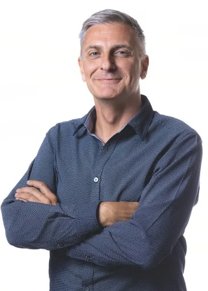 Homme souriant, bras croisés, portant une chemise bleue à motifs sur fond blanc.
