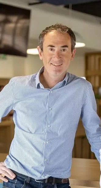 Homme souriant en chemise rayée bleue, posé dans un intérieur lumineux et moderne.
