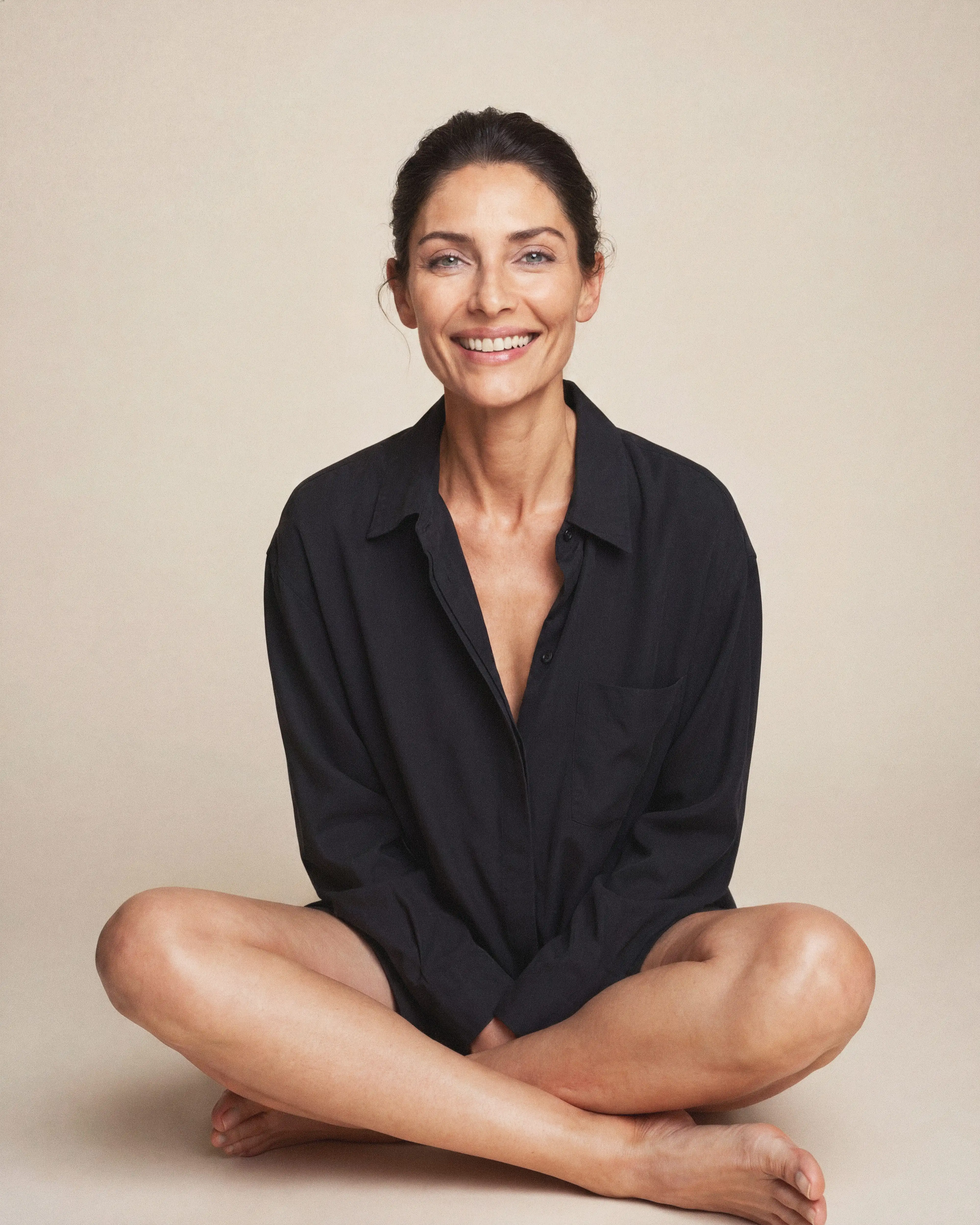 A smiling woman with brown dark hair is smiling while sitting with crossed legs on the floor with a beige backround.