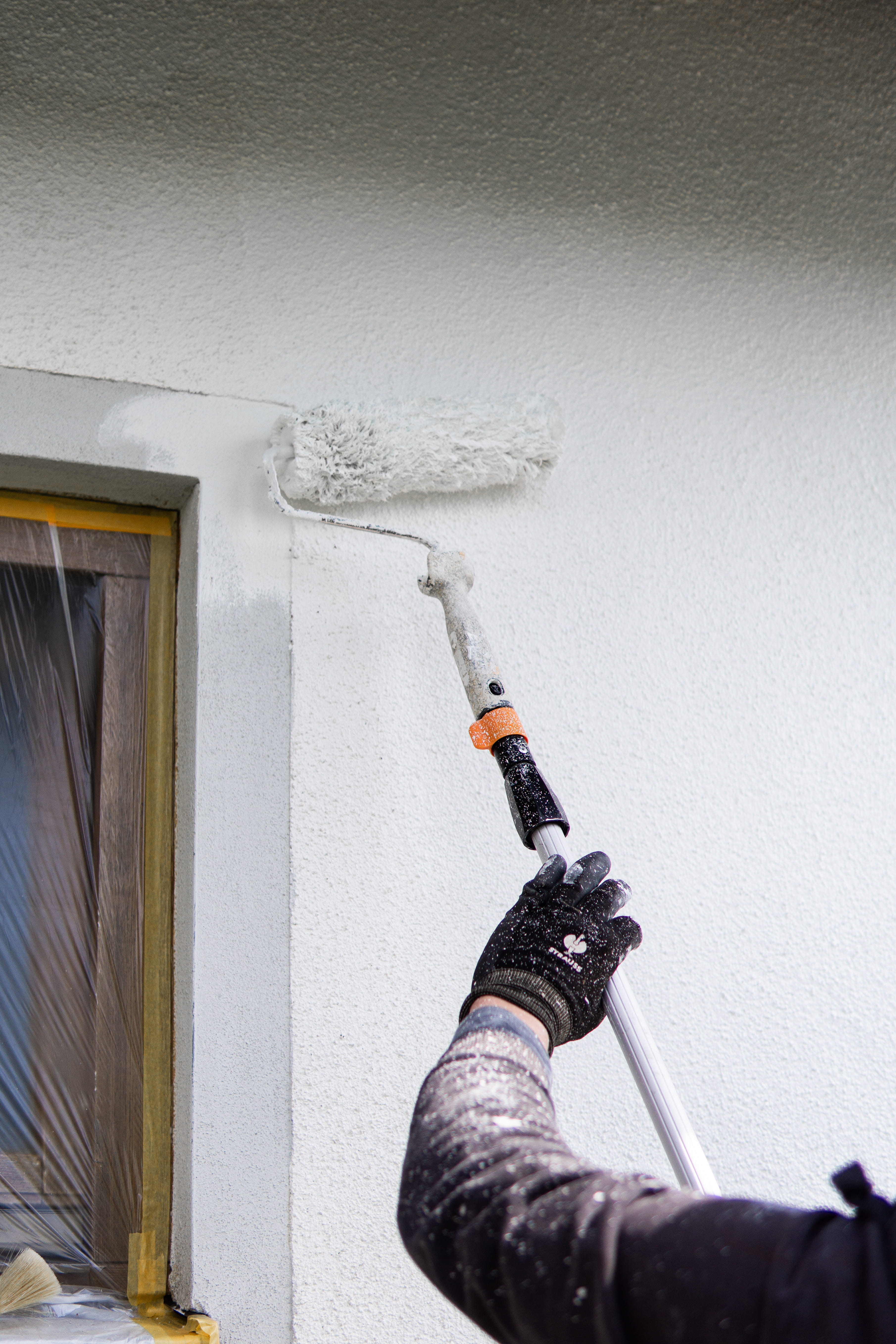 Person streicht mit einer Farbrolle an einer Verlängerungsstange eine weiße, strukturierte Außenwand neben einem Fenster.