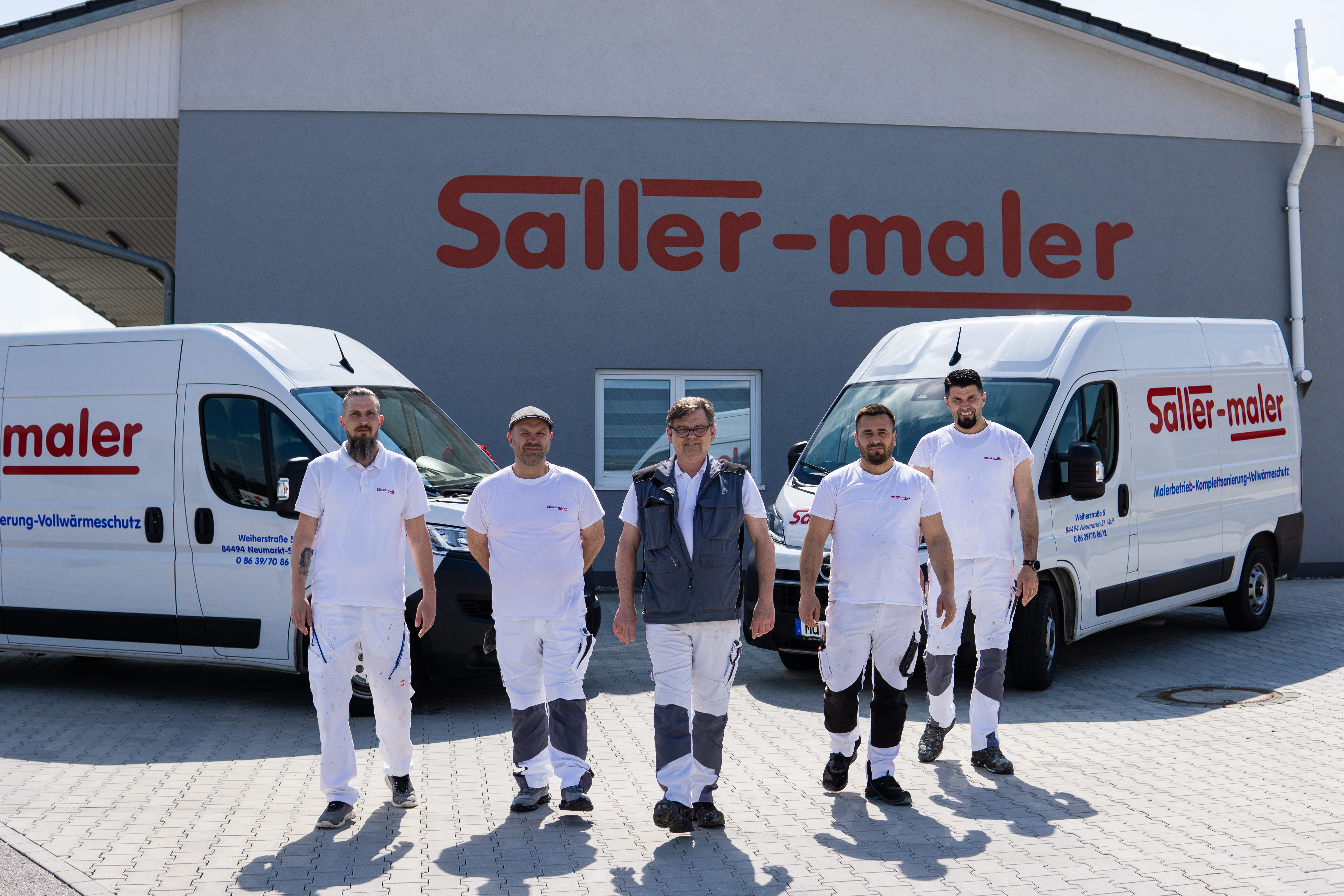 Fünf Maler gehen vor einem grauen Gebäude mit zwei weißen Firmentransportern mit Saller-Maler-Beschriftung.