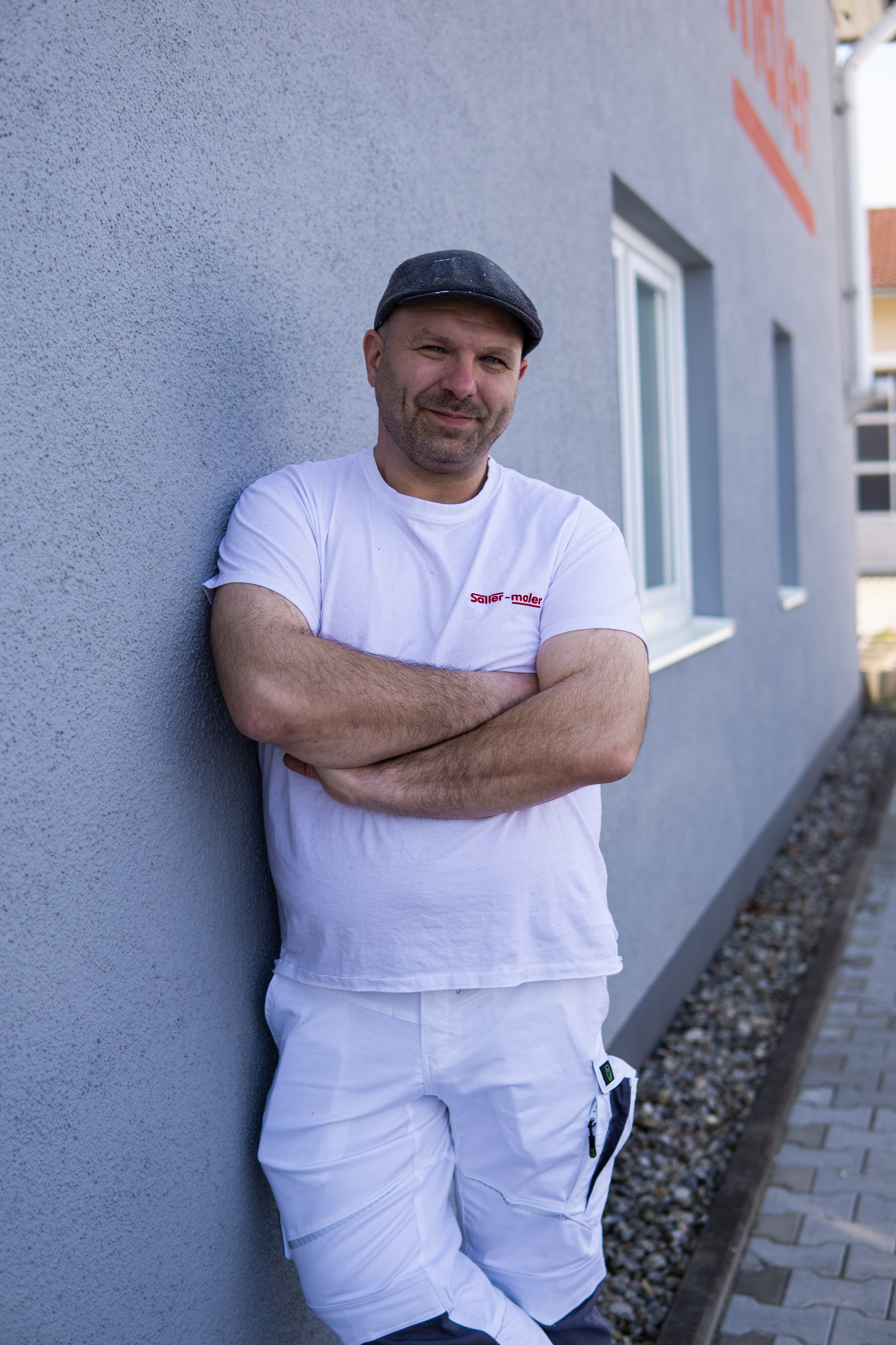 Mann in weißer Malerkleidung mit Mütze lehnt mit verschränkten Armen an einer blaugrauen Wand.