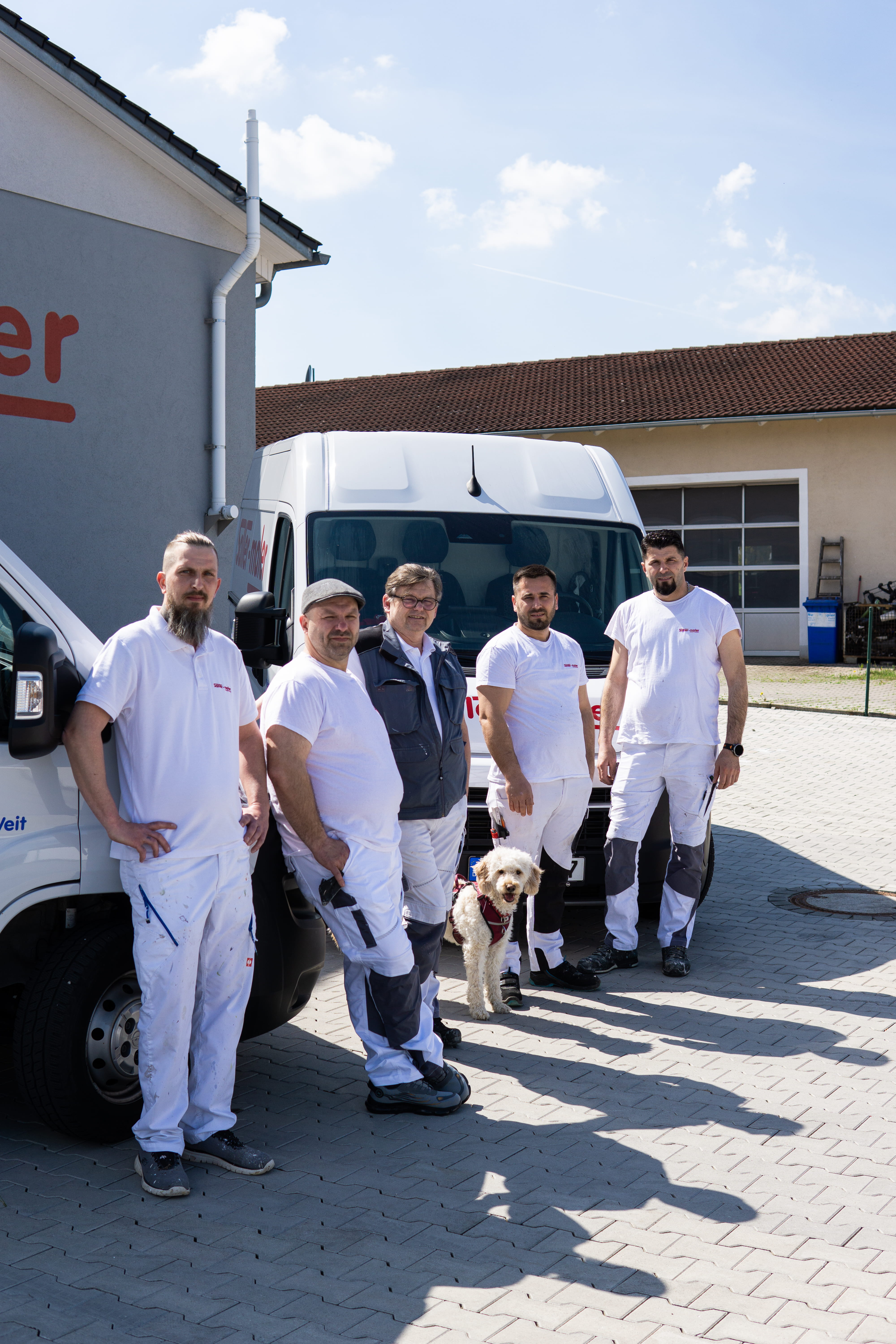 Fünf Männer in Arbeitskleidung und ein Hund stehen im Freien vor zwei weißen Transportern.