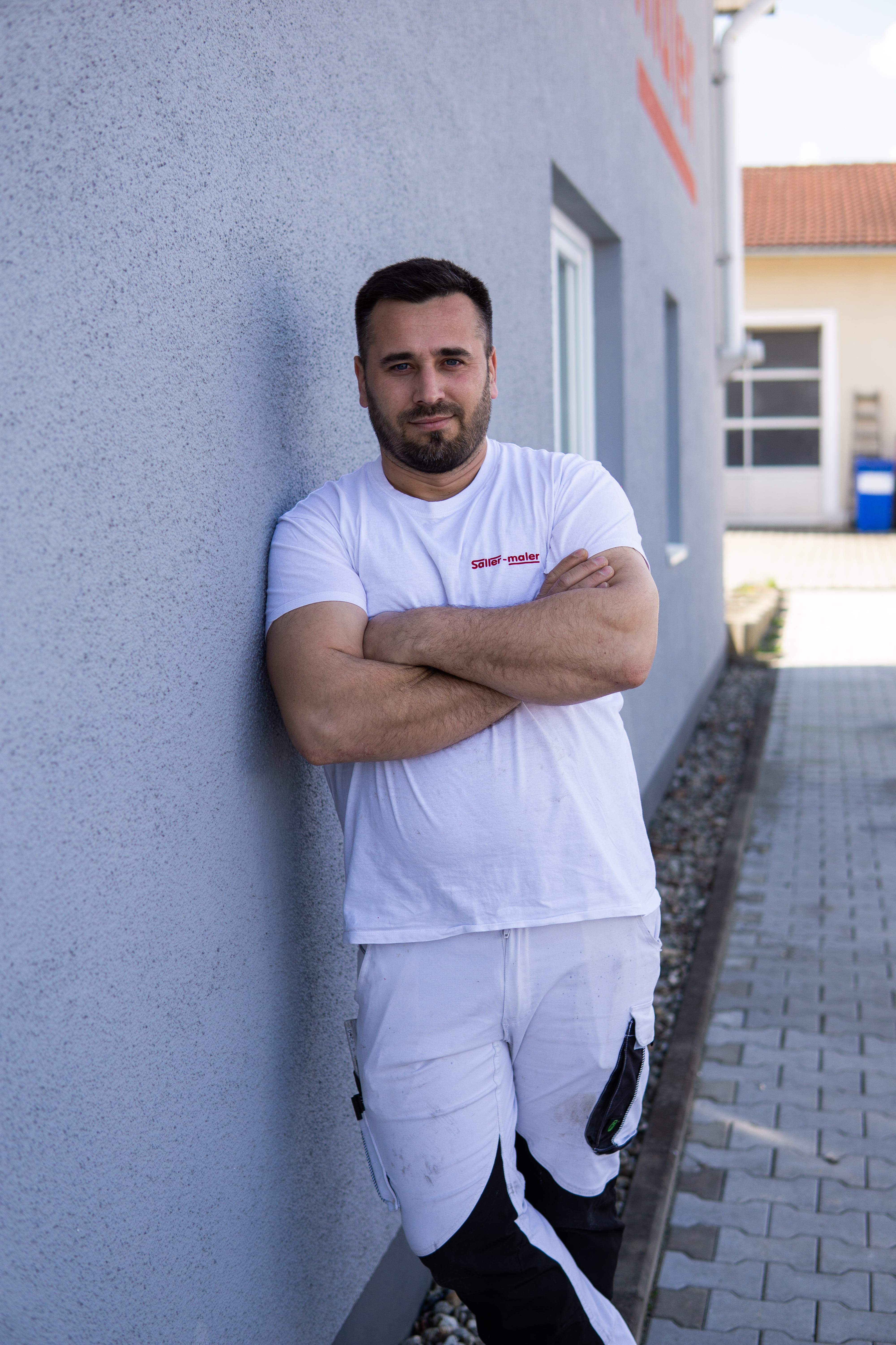 Mann mit verschränkten Armen lehnt an einem sonnigen Tag an einer grauen Hauswand.