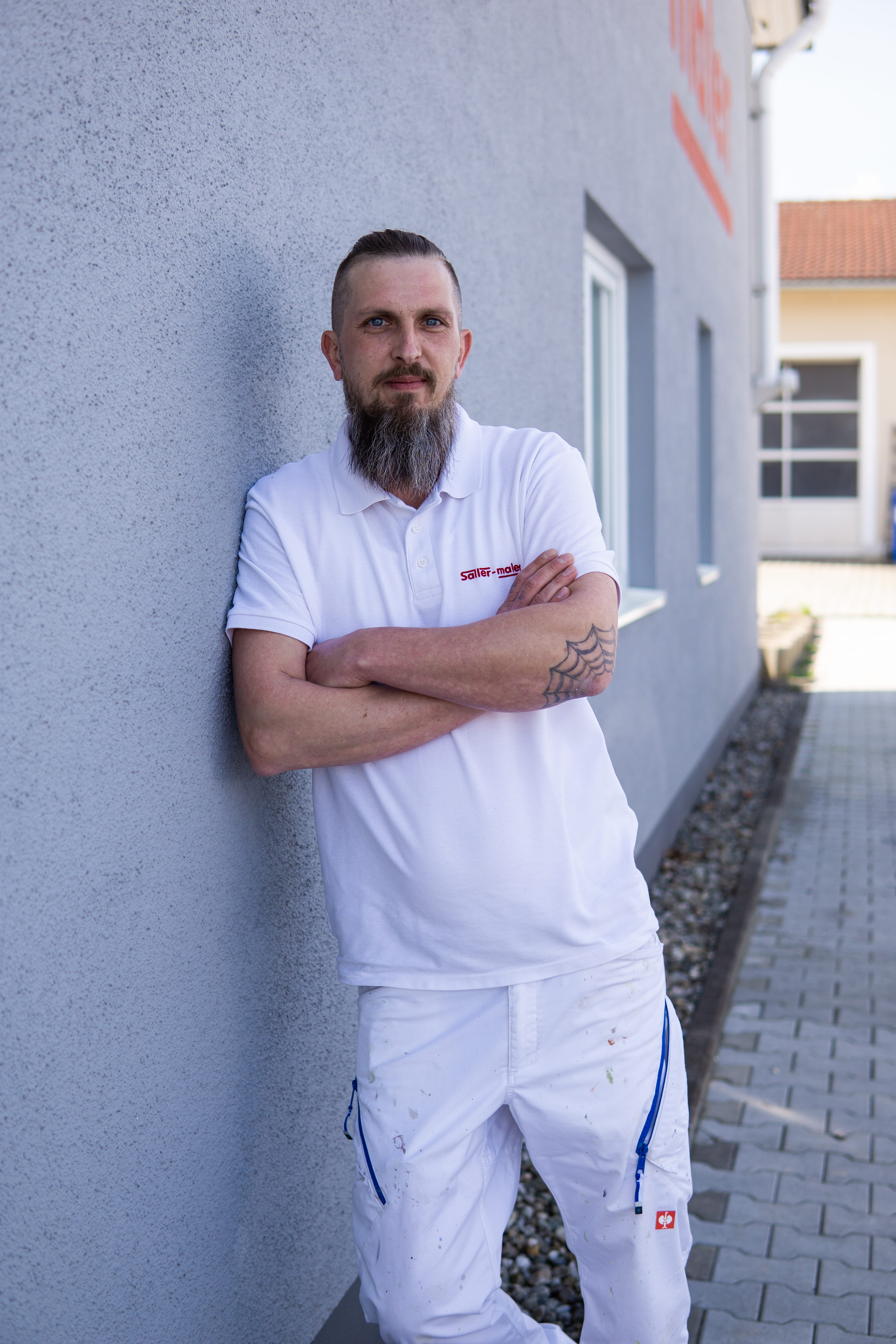 Mann mit Bart und Tattoo in weißer Malerkleidung lehnt draußen an einer grauen Wand.