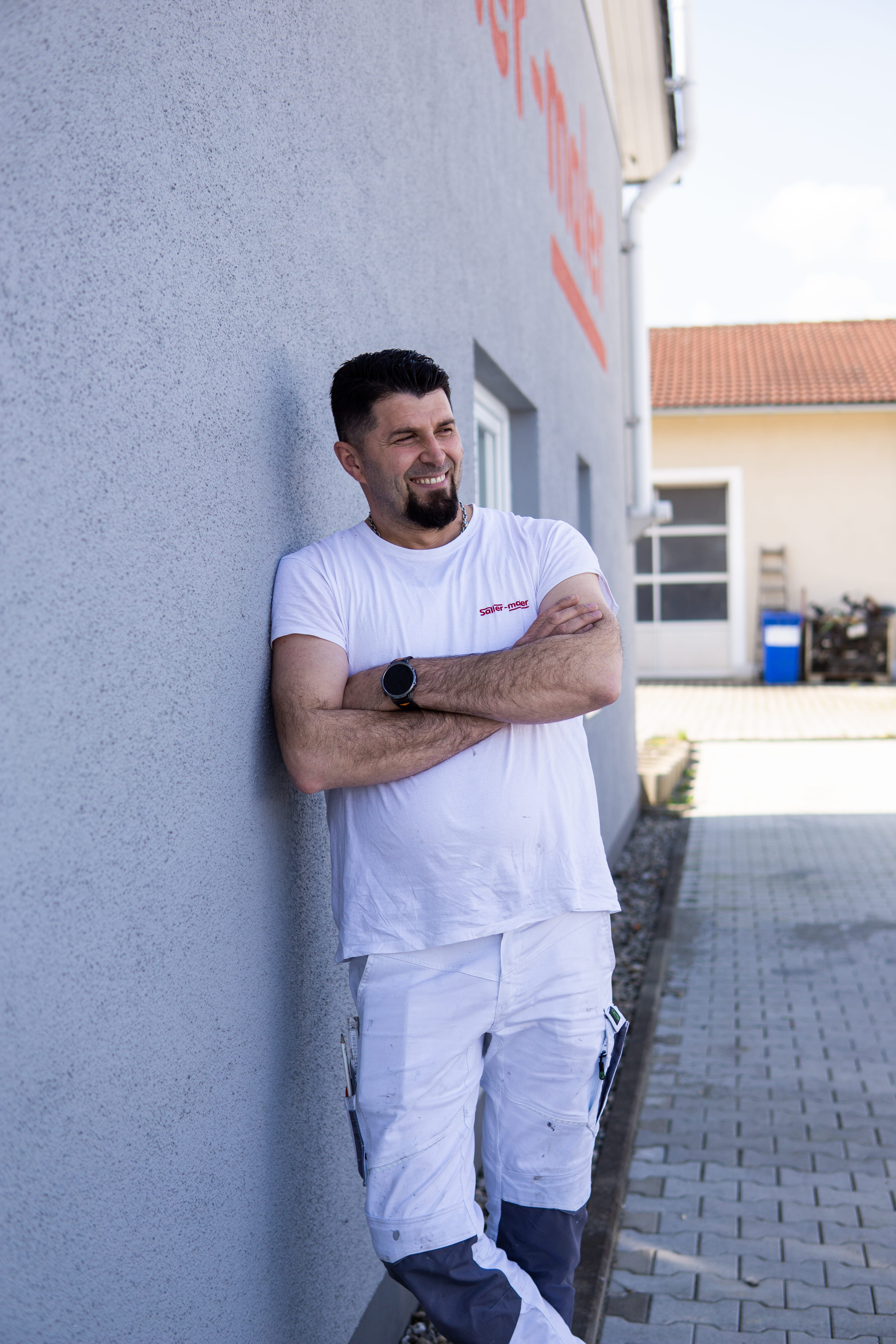 Lächelnder Mann in weißer Arbeitskleidung lehnt mit verschränkten Armen an einer grauen Hauswand.