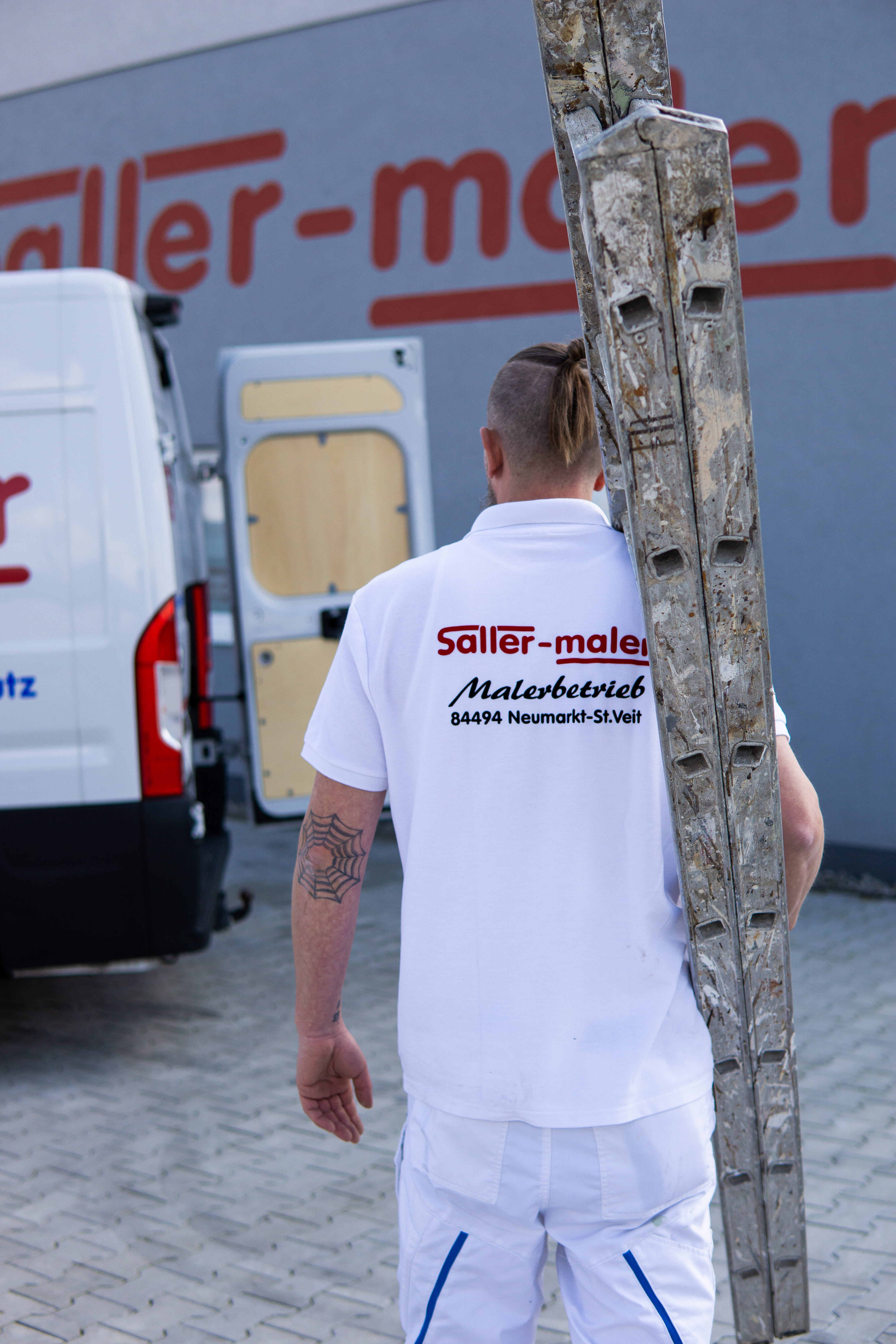 Mann in weißer Arbeitskleidung trägt eine farbbespritzte Leiter auf der Schulter neben einem weißen Transporter.