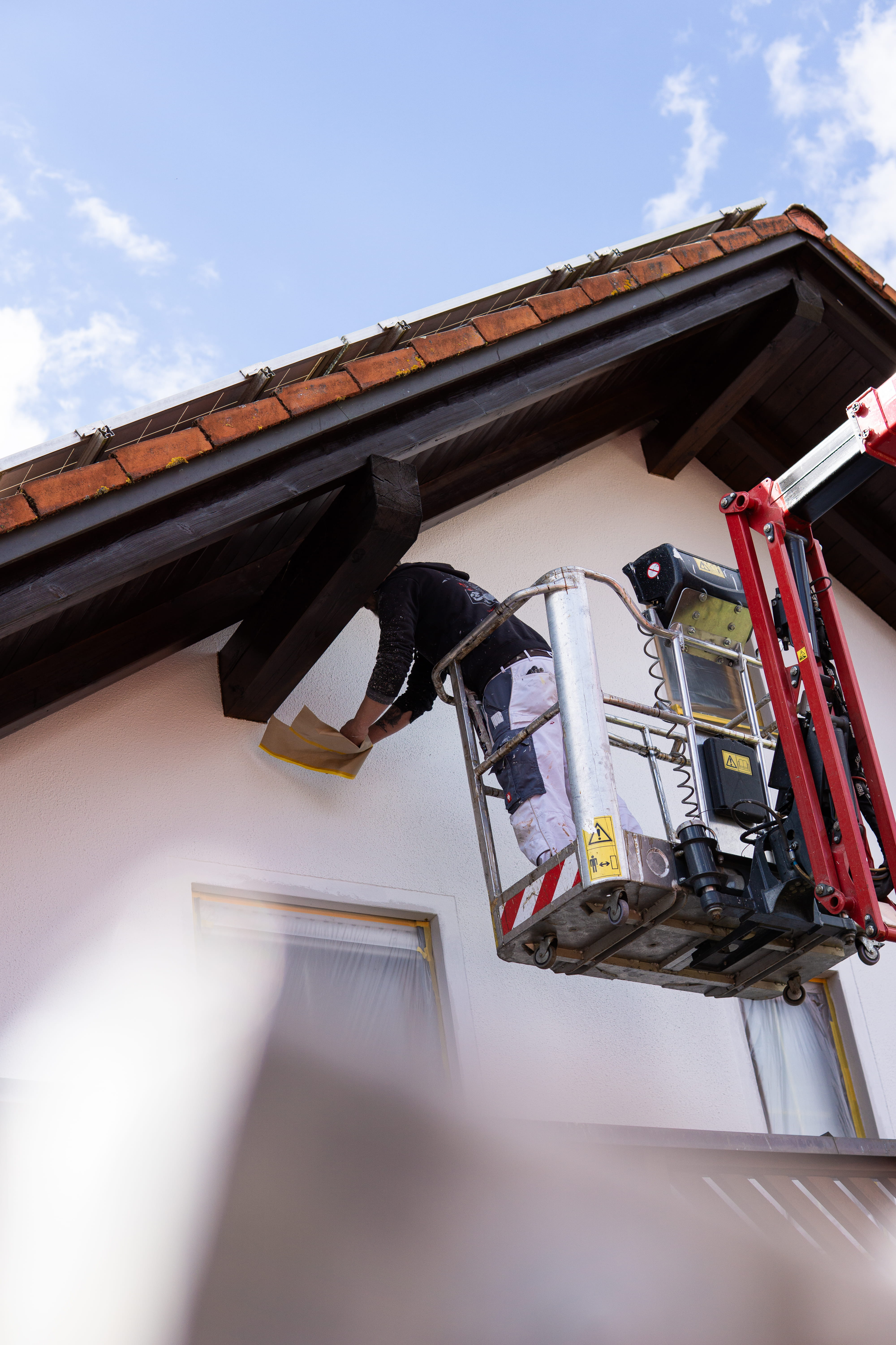 Arbeiter auf einer Hebebühne streicht die obere Außenwand nahe dem Hausdach mit Solarmodulen.