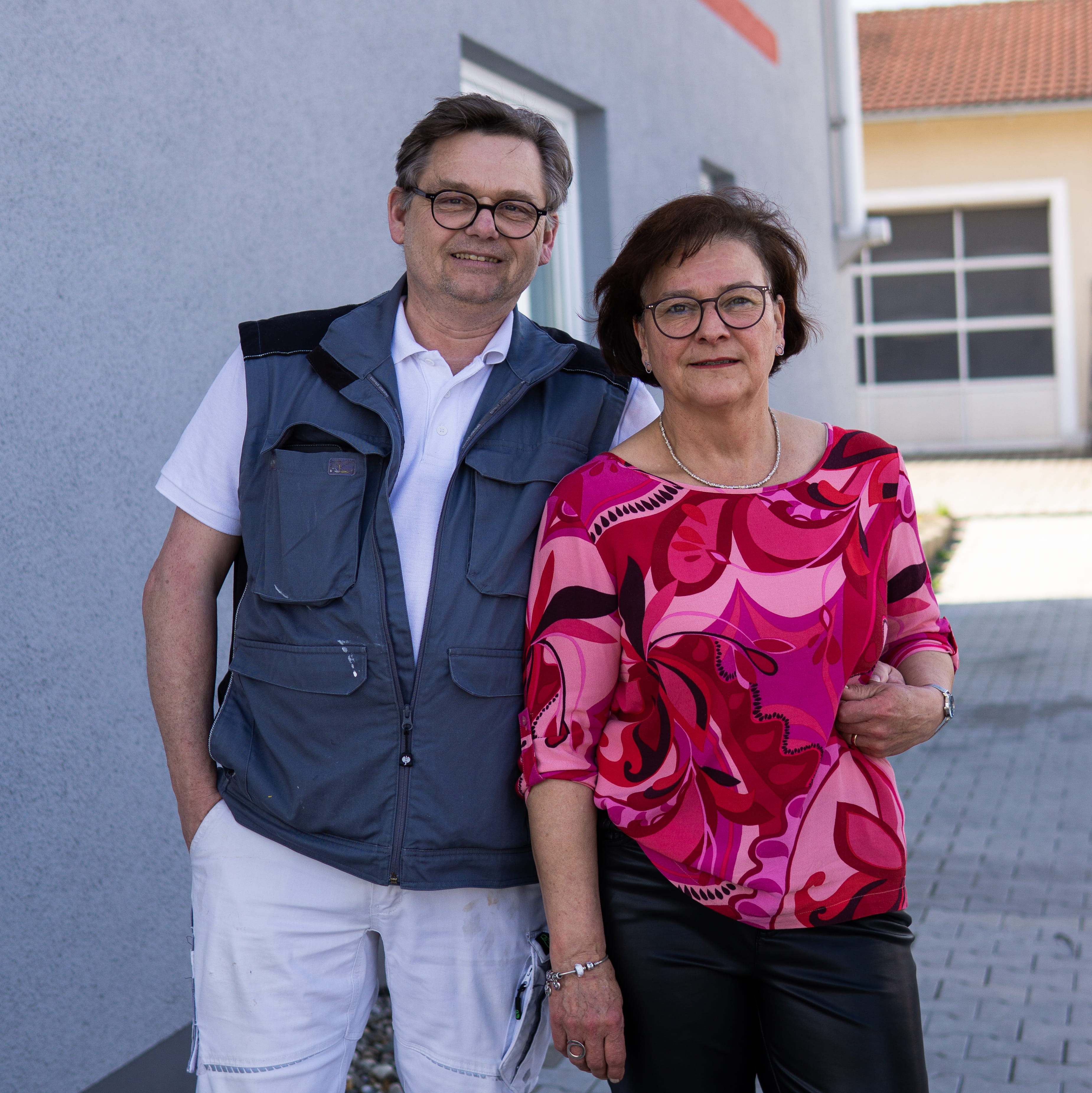 Smiling man with glasses in workwear standing against a gray building wall with arms crossed.
