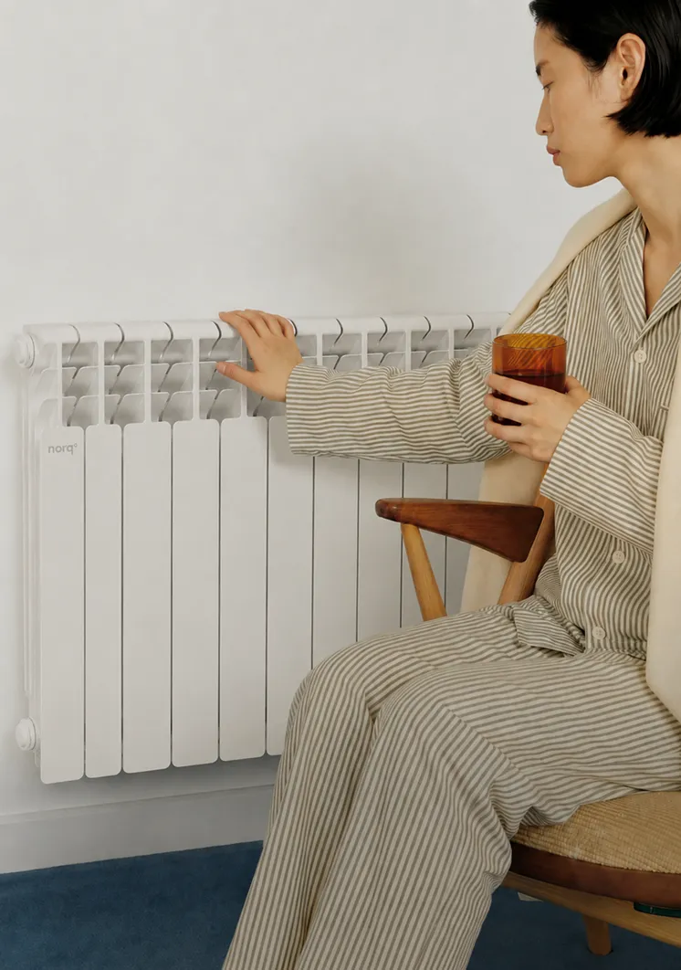 Person in striped pajamas sitting and touching a white wall-mounted radiator while holding a brown glass.