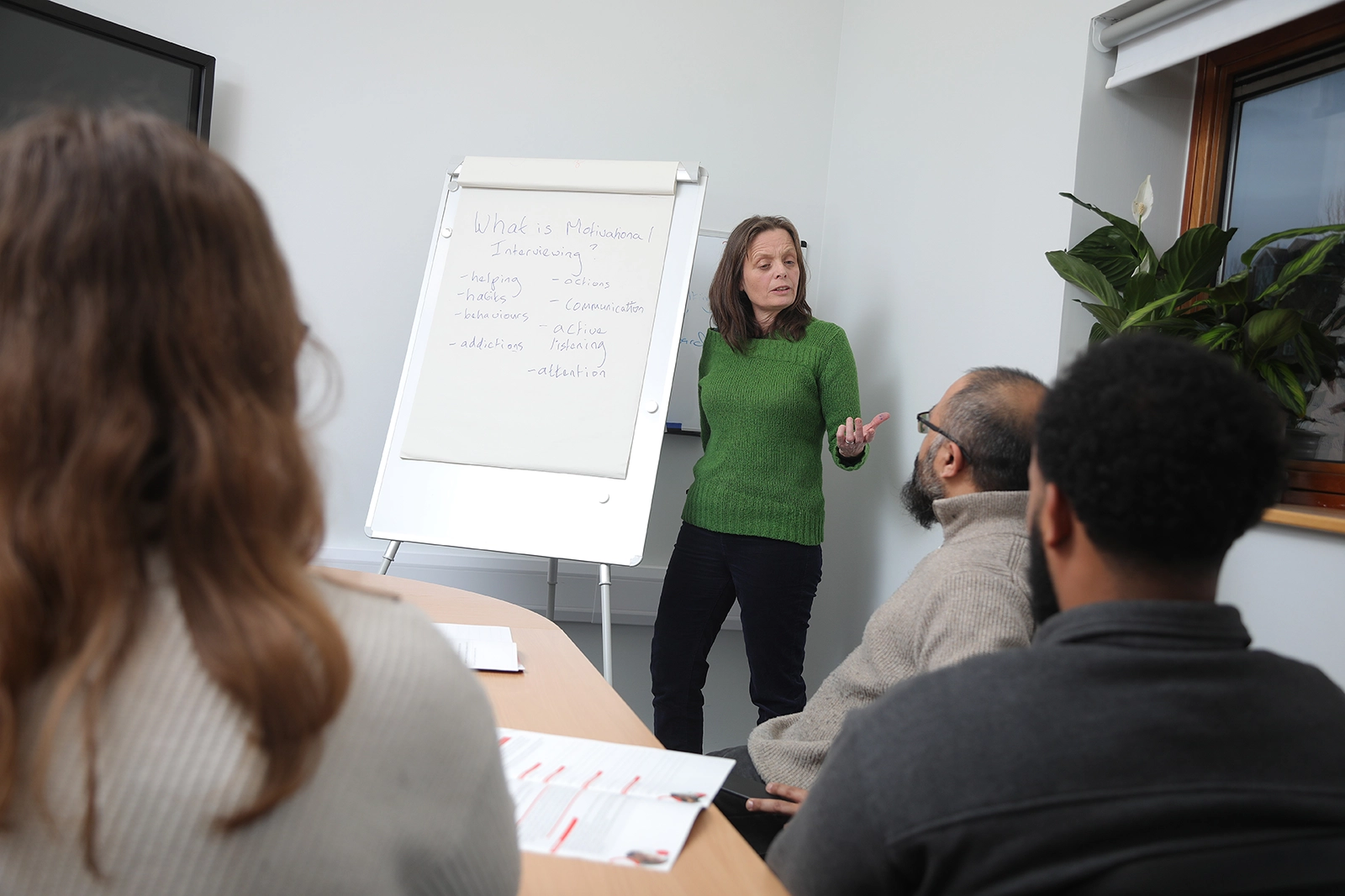 Woman in green jumper presenting motivation interviewing on flipchart to seated group in meeting room.