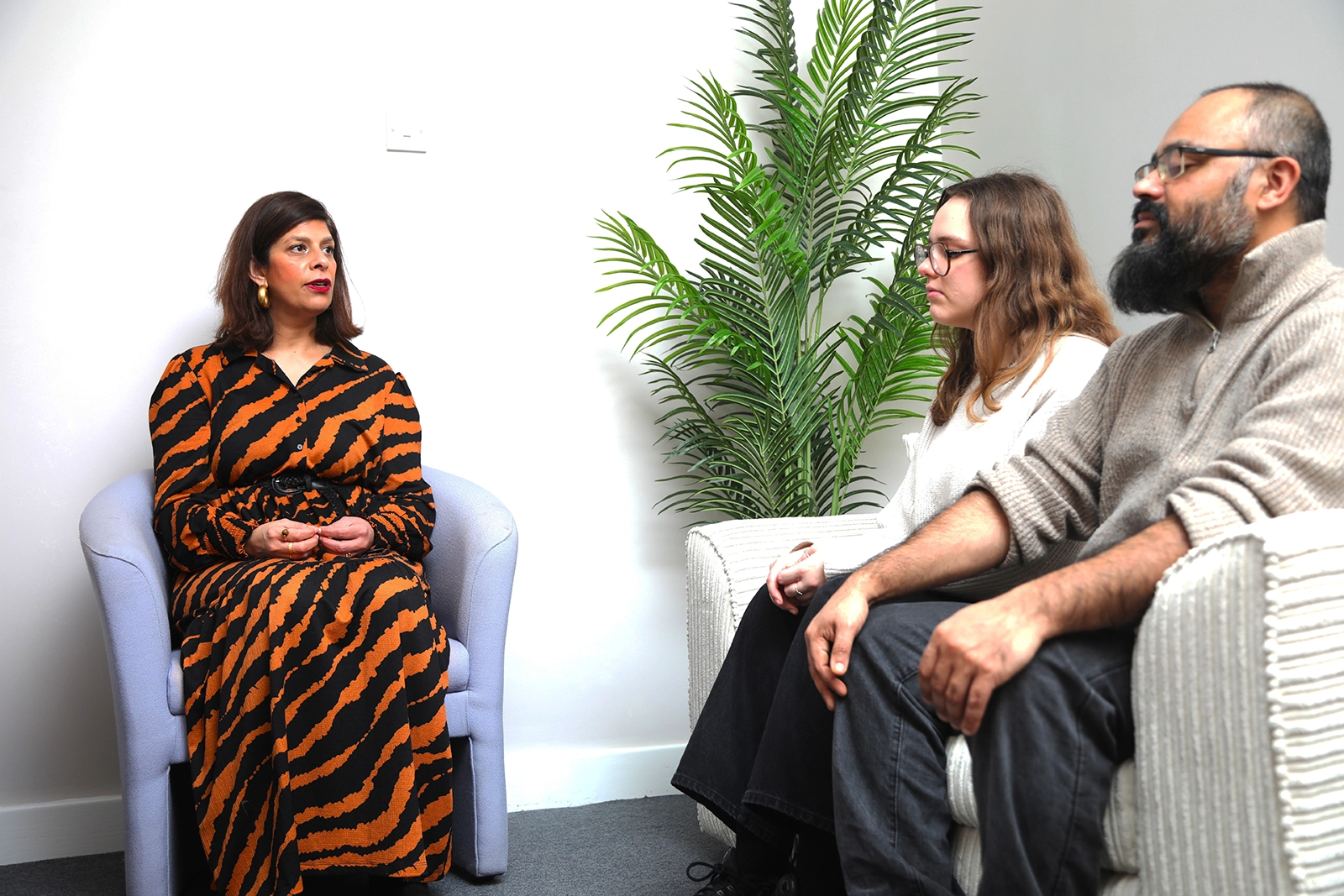 Woman in tiger stripe dress seated opposite a man and woman sitting on a couch next to a green plant.