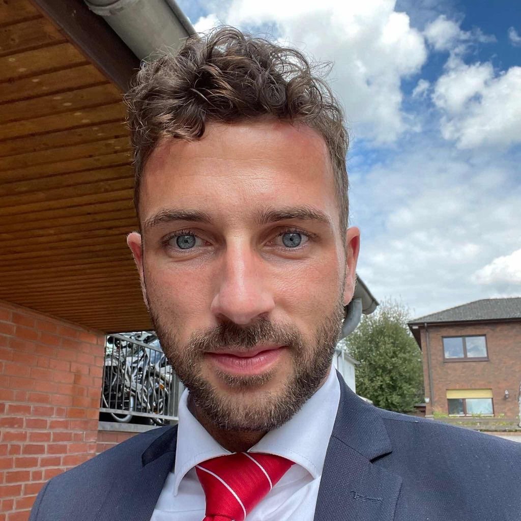 Man with curly hair and beard wearing a suit and red tie outside near brick buildings under blue sky.