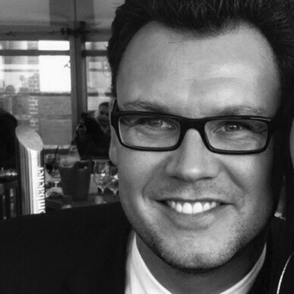 Close-up of a smiling man wearing glasses in a warmly lit indoor setting.