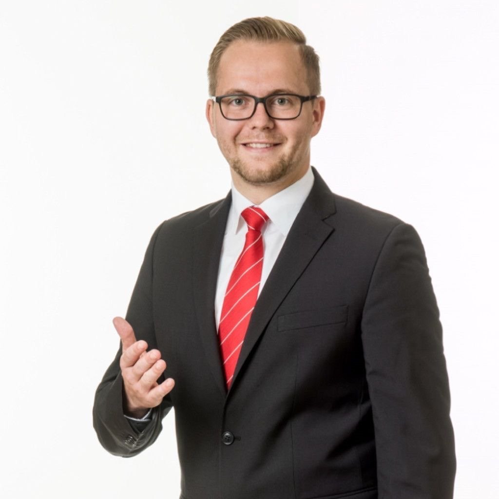 Man with glasses wearing black suit, white shirt, and red striped tie gesturing with his right hand.